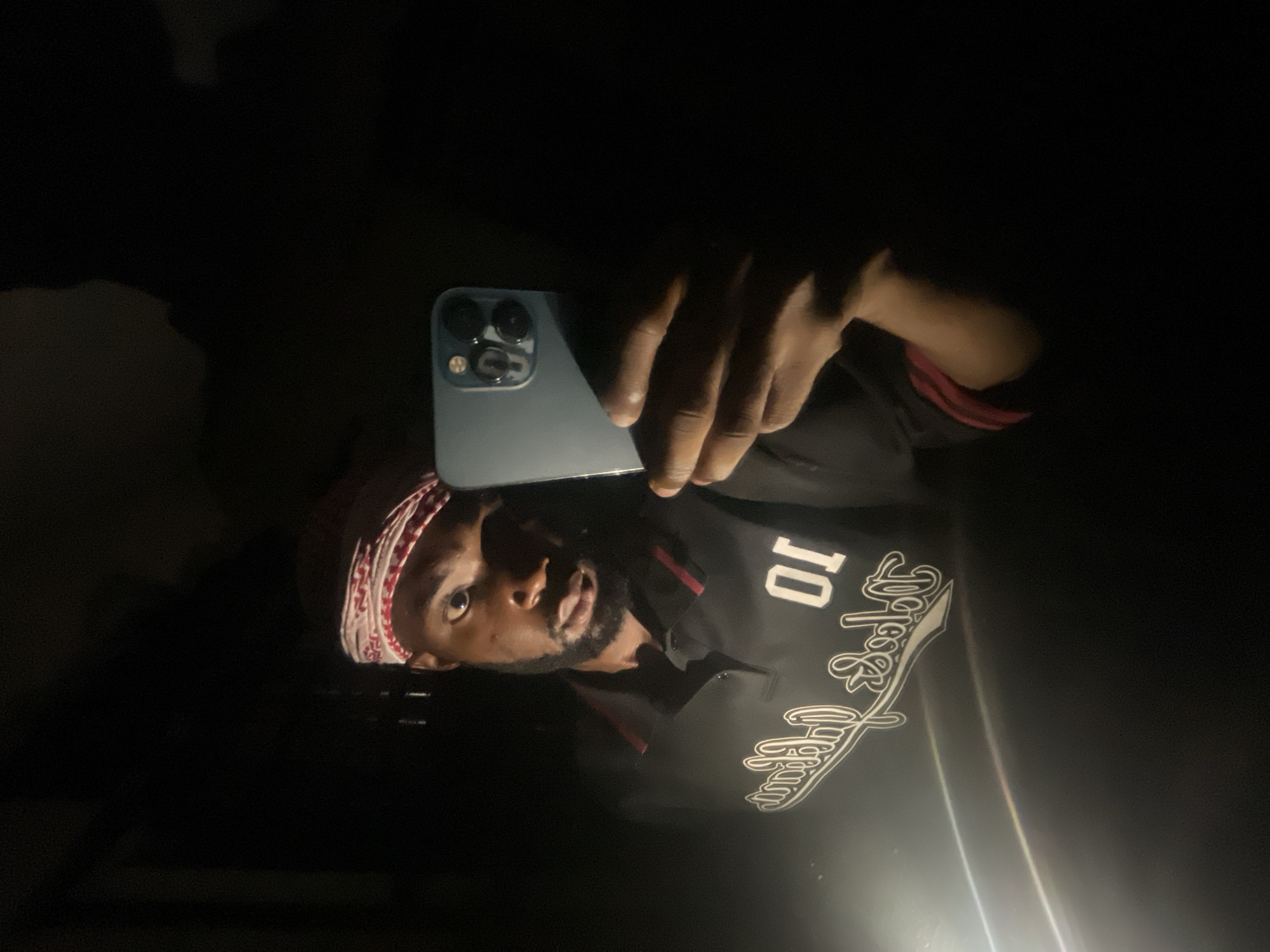 A man is taking a selfie with a smartphone. He is wearing a black shirt with the number 10 and a white and red patterned head covering. The environment is dark, with only a few light sources illuminating the man and the phone. The location is Lokoja, Nigeria.