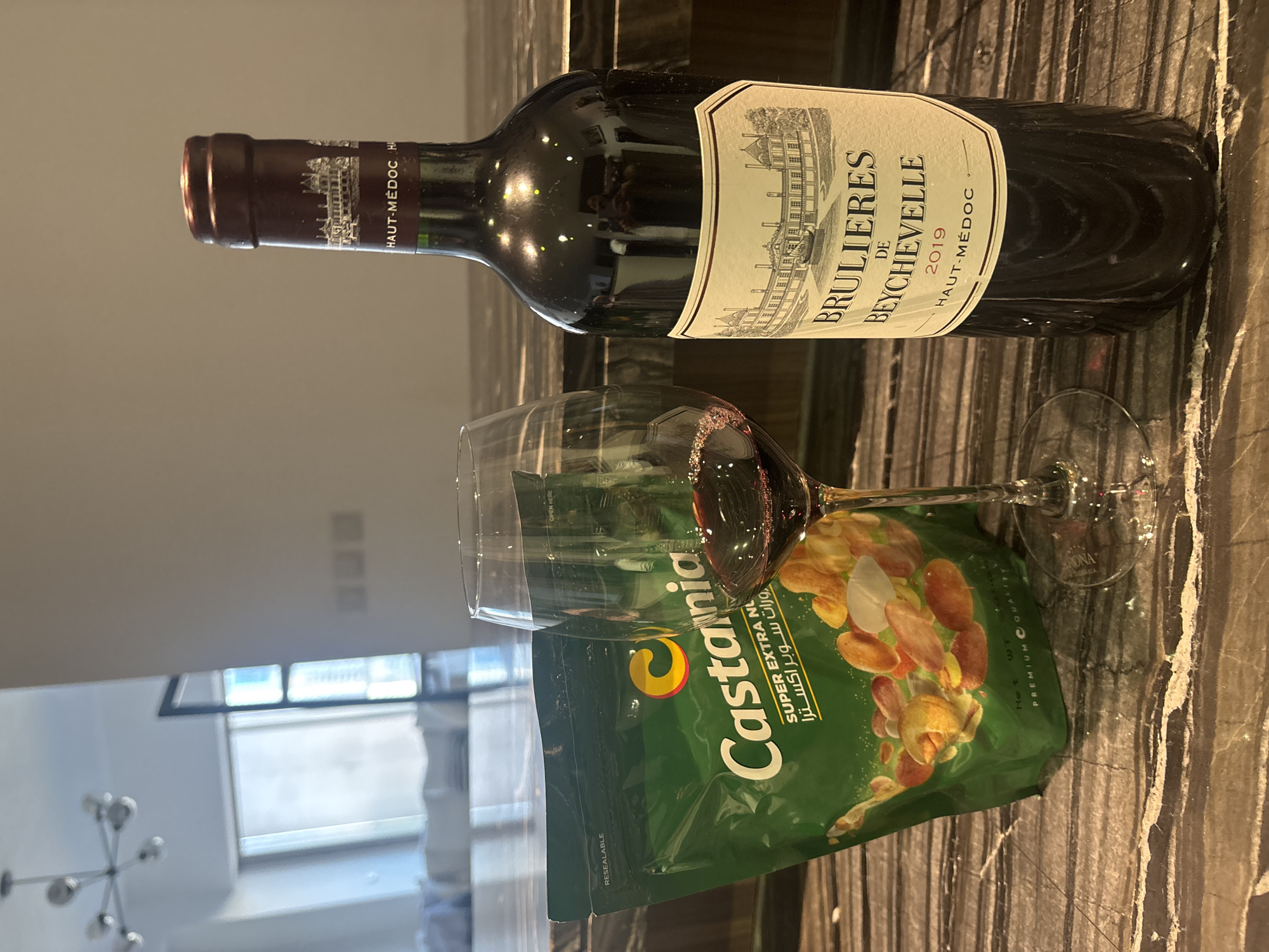An indoor, eye-level, medium shot depicts a still life arrangement of a bottle of red wine and a bag of nuts next to a wine glass. The bottle of red wine is standing upright and is mostly in focus. It is dark red with a burgundy label that has a white illustration of a chateau and text that reads "BRULIERES DE BEYCHEVELLE 2019 HAUT-MEDOC". The bag of nuts is green and also mostly in focus. It has a large yellow "C" logo and the word "Castania" printed on it, along with text that reads "SUPER EXTRA NUTS" in English and Arabic. A wine glass filled with red wine is placed between the bottle and the bag of nuts. The objects are resting on a speckled, dark brown marble countertop. In the background, out of focus, is a bright, modern interior with a light fixture hanging from the ceiling and a window with a view of a city skyline. The lighting is bright and artificial, suggesting an indoor setting in an apartment or hotel. The scene is likely in Dubai, United Arab Emirates, given the presence of Arabic script on the nut packaging and the modern, high-rise architectural style visible in the background. No people are visible in the frame.