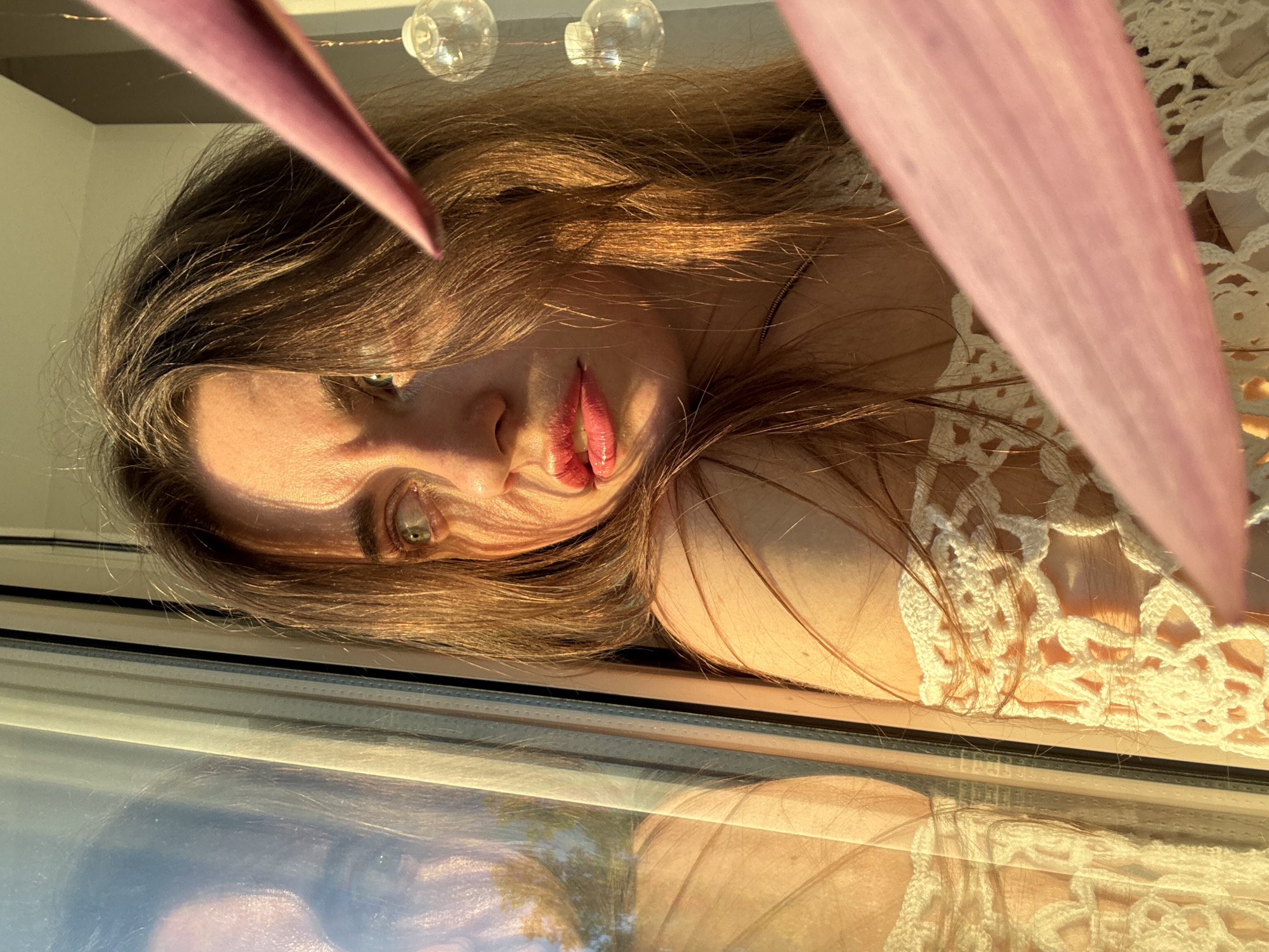 This media file captures a close-up portrait of a woman lying horizontally, illuminated by warm, golden light, suggestive of late afternoon or sunset in Chaikovsky, Russia. Her face, framed by long, brown hair, is prominent, and she gazes directly at the viewer with a focused, perhaps pensive, expression. Her lips are slightly parted, revealing red lipstick, and her light-colored eyes stand out.
A large, blurred pink or purple leaf or plant part diagonally crosses the upper right foreground. In the background, above her head, two clear, spherical decorative lights are faintly visible. The woman is wearing a white, open-knit or crocheted top.
The bottom portion of the image shows a reflective surface, likely a window, where a blurred reflection of her form and indistinct outdoor elements, possibly trees and sky, can be discerned. The strong directional light highlights the textures of her hair and clothing, creating an intimate and warm atmosphere. No text is visible.