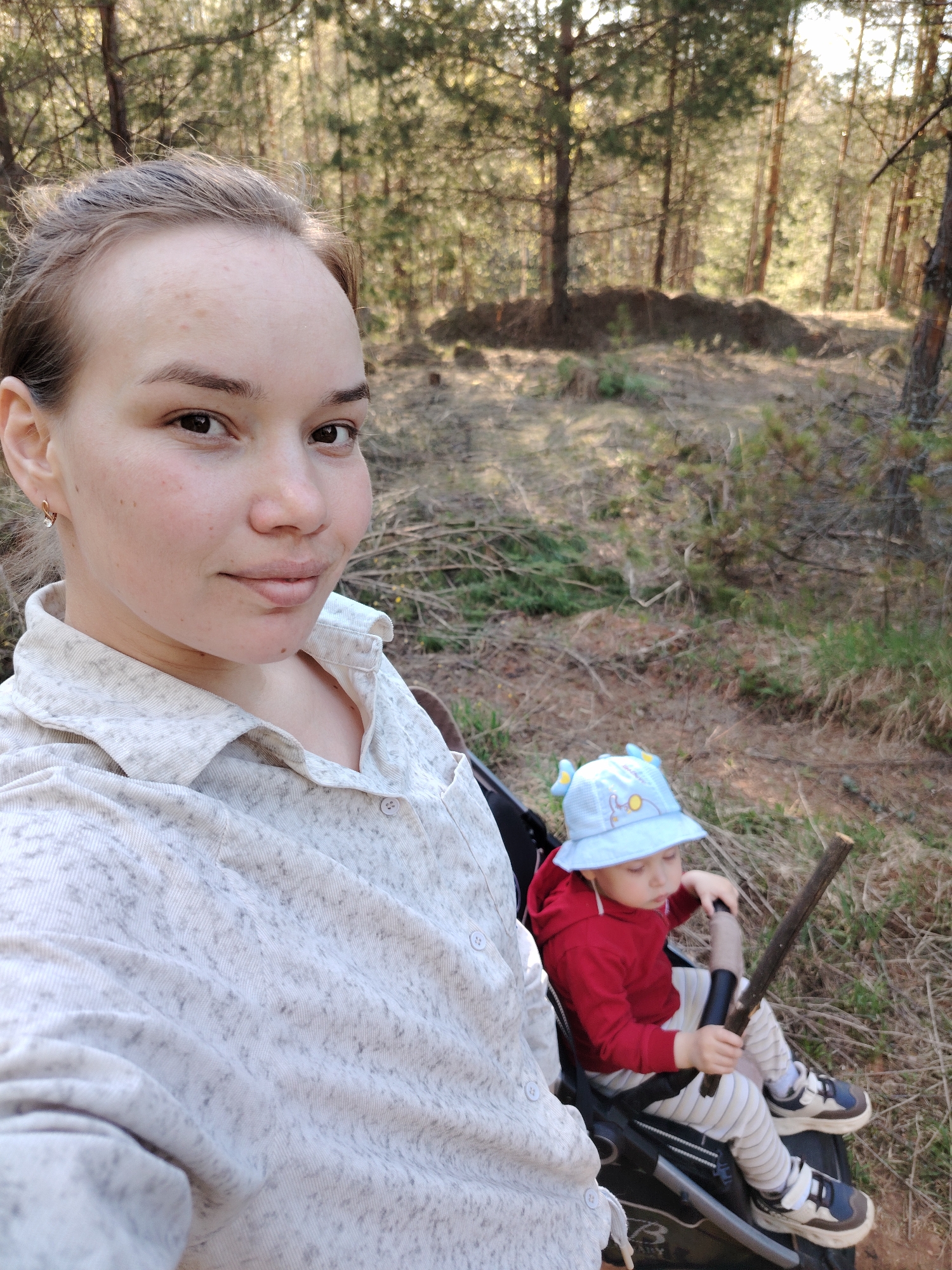 In a daytime, outdoor scene, a woman and child are positioned in the vicinity of Марковский, Russia, within a forest environment characterized by slender trees, possibly pine or similar coniferous species. The woman, facing the camera, has fair skin and her hair is pulled back. She is wearing a light-colored, button-down shirt. A child, seated in a stroller, is wearing a red sweatshirt, striped pants, and sneakers, along with a blue sun hat. The child is holding a stick. The surrounding area contains fallen branches and dense foliage. The lighting is bright, suggesting a sunny day.