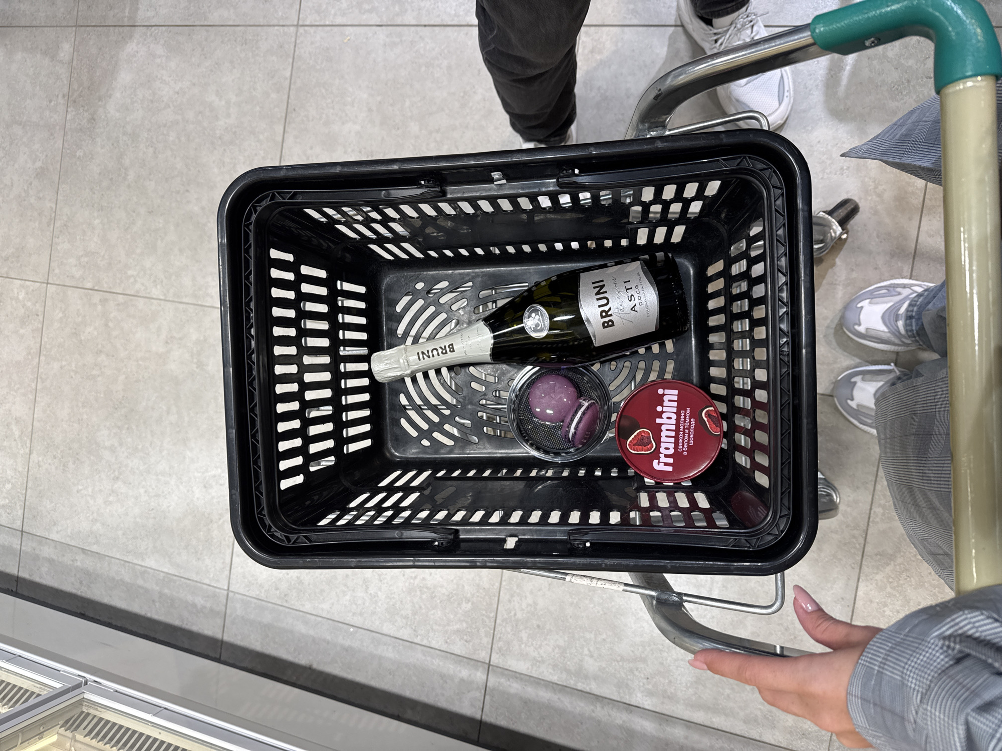 Captured from an overhead perspective, the image focuses on a black plastic shopping basket situated within a shopping cart. Inside the basket, a bottle of Bruni Asti D.O.C.G. wine is visible, accompanied by a container of Frambini chocolate and a glass holding macarons. The cart is held by a hand with light pink nails and light skin. This scene occurs indoors, likely in a supermarket or a store. The location remains Unknown, Unknown.