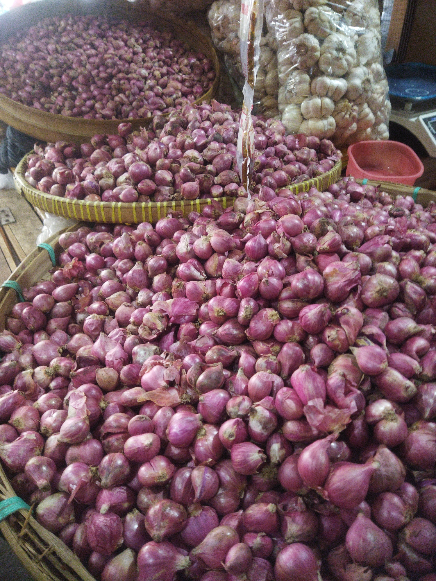 The image displays a market stall in Wonogiri, Indonesia, overflowing with a generous amount of red onions. The produce is arranged in woven baskets and large piles, showcasing their vibrant purple hue and varying sizes. Some of the onions are still connected in clusters, while others are loose. In the background, a large bag filled with garlic cloves is visible, suggesting a broader selection of produce available. The overall scene depicts a bustling marketplace, rich in local ingredients.