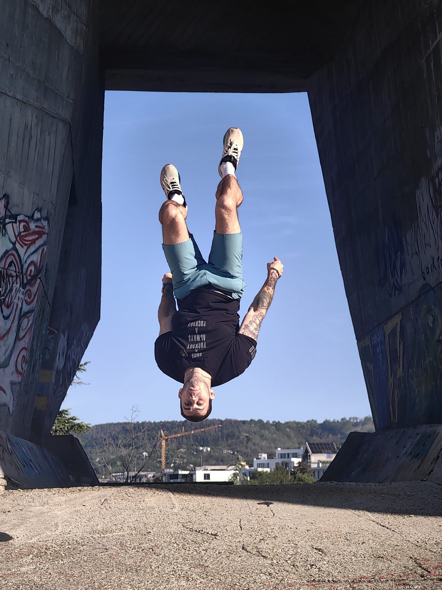 A man is upside down, performing a backflip under a concrete structure. The concrete structure appears to be part of a bridge or overpass, with graffiti visible on its surfaces. The background reveals a cityscape with buildings, hills, and a clear blue sky. A construction crane is visible. The ground is a rough, gravel-like surface. Based on the image, the location is likely in or near Varna, Bulgaria, due to the architectural style and surrounding landscape.