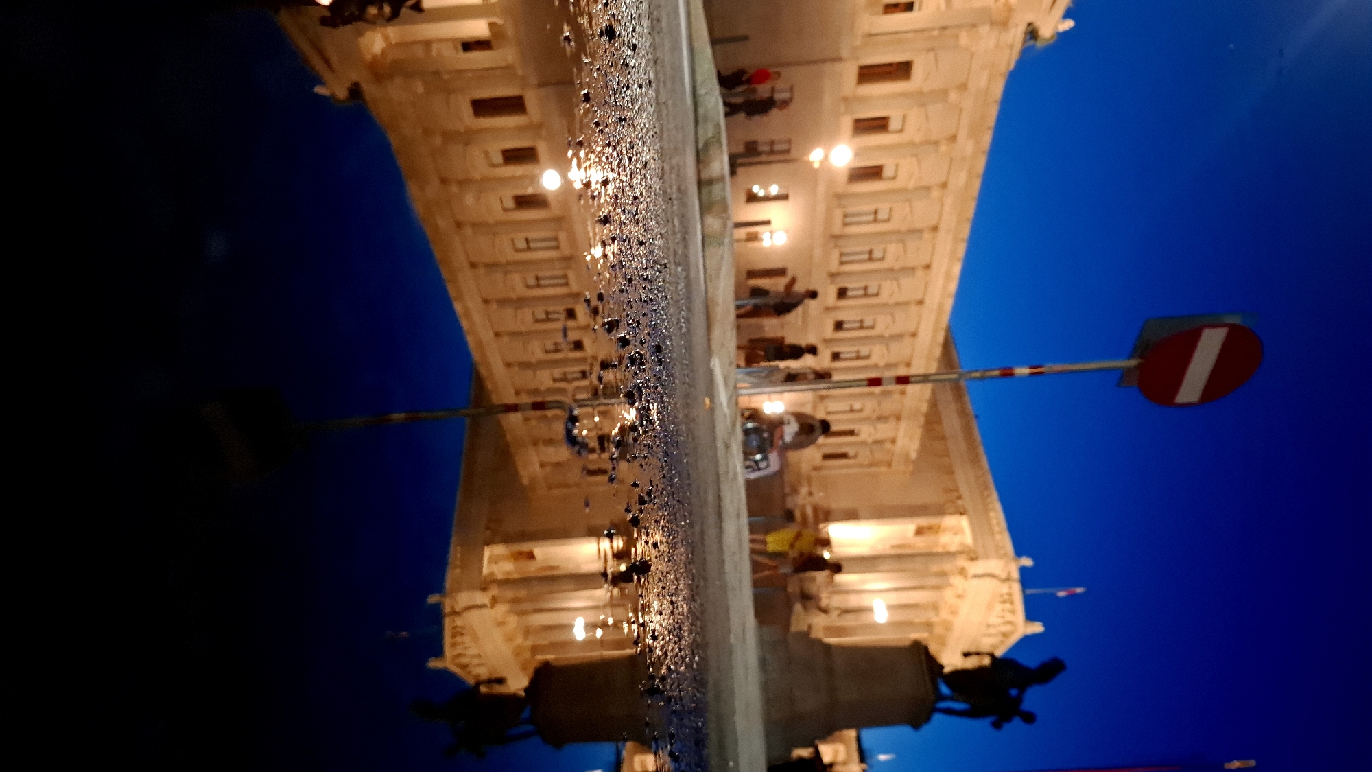 The image depicts the exterior of the Austrian Parliament Building in Vienna, Austria, at dusk. The building is illuminated, casting a warm glow on its classical facade. In the foreground, a wet pavement reflects the building and the clear, deep blue sky. A no-entry traffic sign on a pole is visible in the mid-ground, slightly to the left of center. A statue of a horse and rider, silhouetted against the sky, stands to the right of the Parliament building. Several individuals are seen walking in front of the building, their figures blurred due to the low light conditions and the photograph's focus on the reflection. The overall scene is one of a tranquil evening in a historic European city.