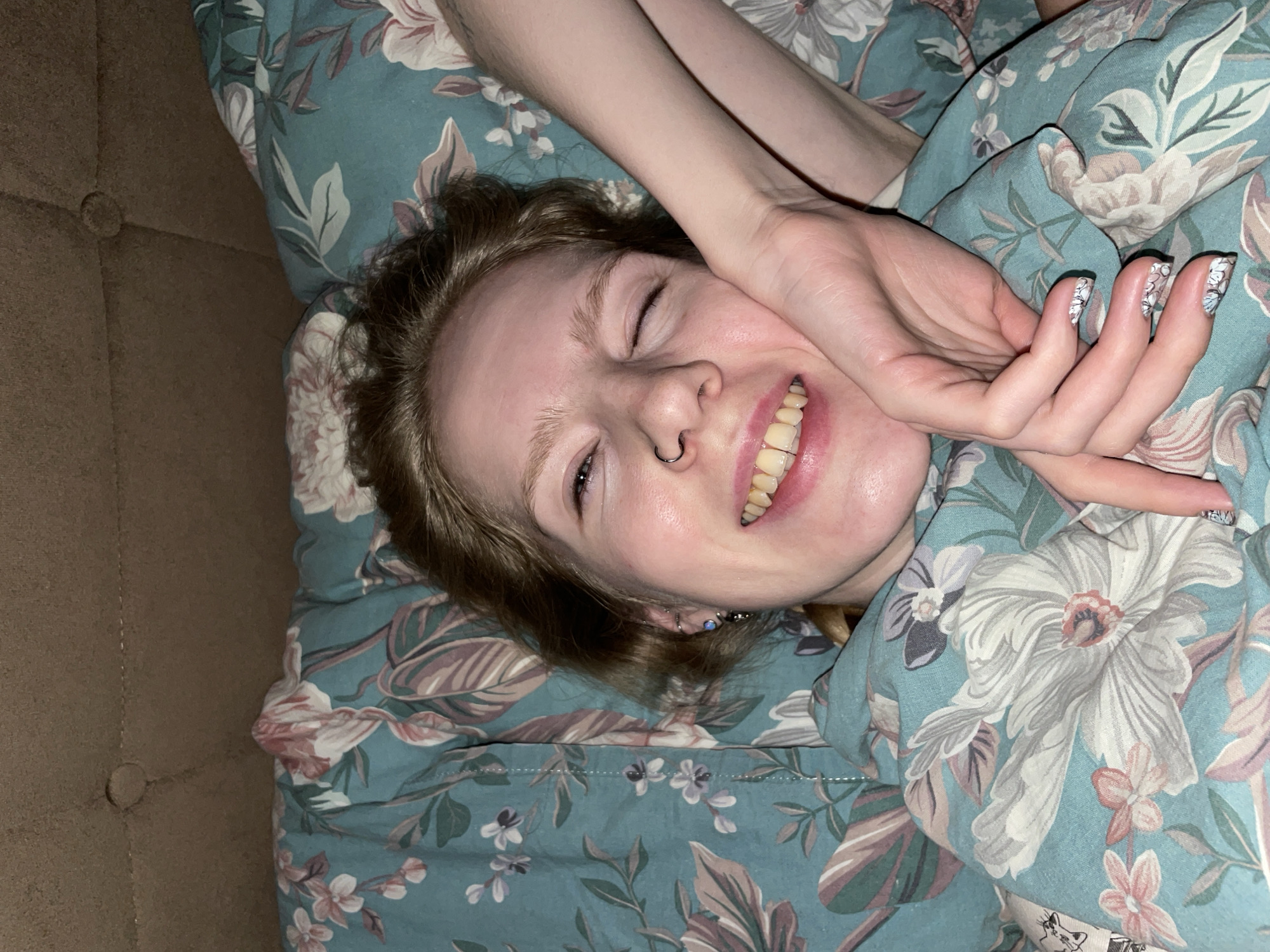A person with light skin, light brown hair, and a nose ring is smiling at the camera. Their eyes are partially closed, and they appear to be lying down. They are covered with a blue floral patterned blanket, and their hands are visible near their face. The left side of the image includes a brown textured surface. The location is not determinable from the image.