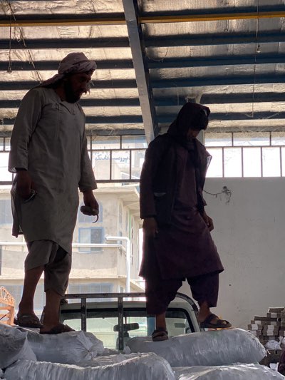 Two men stand on top of bags loaded onto a white truck. The man on the left wears a head covering, a long tunic, and sandals, holding an object in his right hand. The man on the right wears a dark robe with a hood and sandals. The background shows a building and a corrugated metal ceiling. A sign in the background contains Farsi script. The scene is located in the city of Shurandam, Afghanistan.