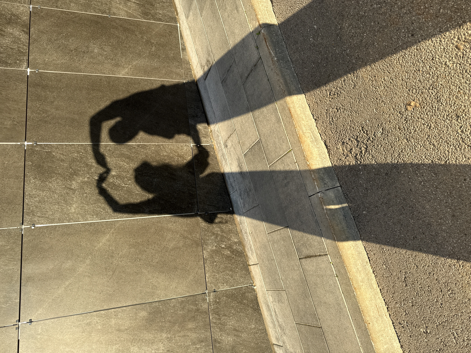 The image captures two shadows of people forming a heart shape on a tiled wall. The shadows are cast from the left side of the frame, indicating the sun's position. The wall is composed of rectangular, dark grey tiles with visible grout lines and metal fasteners. Below the wall is a sidewalk paved with rectangular, light grey tiles. A curb separates the sidewalk from a rough-textured asphalt surface on the right. The lighting suggests it is daytime, likely late afternoon or early morning due to the long shadows. The scene does not provide any specific geographical markers to pinpoint a location within Kazan, Russia.