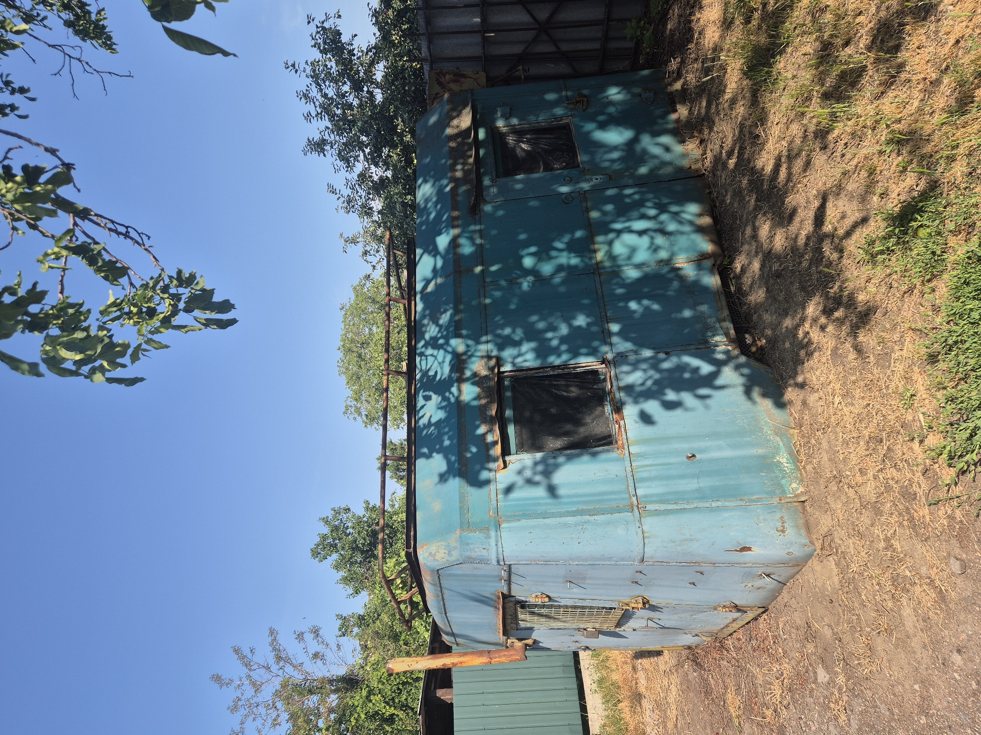 In Sozonivka, Ukraine, an aged, teal-colored metal trailer sits outdoors on a dirt and dry grass area under a clear blue sky. The trailer, with visible rust and signs of wear, has a roof rack. Two windows, one with a metal grate and padlock, are visible on the side. Tree branches with green leaves cast shadows across the trailer and the ground. A tall, rusted pipe extends vertically from the left side of the trailer. In the background, a green fence and a grey metal fence are present, along with lush green trees.