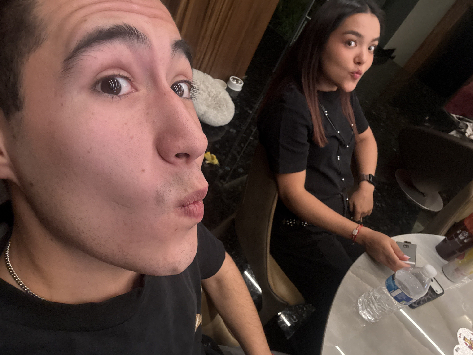 A close-up indoor photograph captures two individuals with pursed lips. The man is in the foreground, partially cropped. He is wearing a dark t-shirt and a silver chain. A woman is seated behind him to his right, also with pursed lips, wearing a dark button-down top. A small table in the lower right corner holds a water bottle and other objects. The location is possibly in a home or restaurant in San Bernardino Tlaxcalancingo, Mexico.