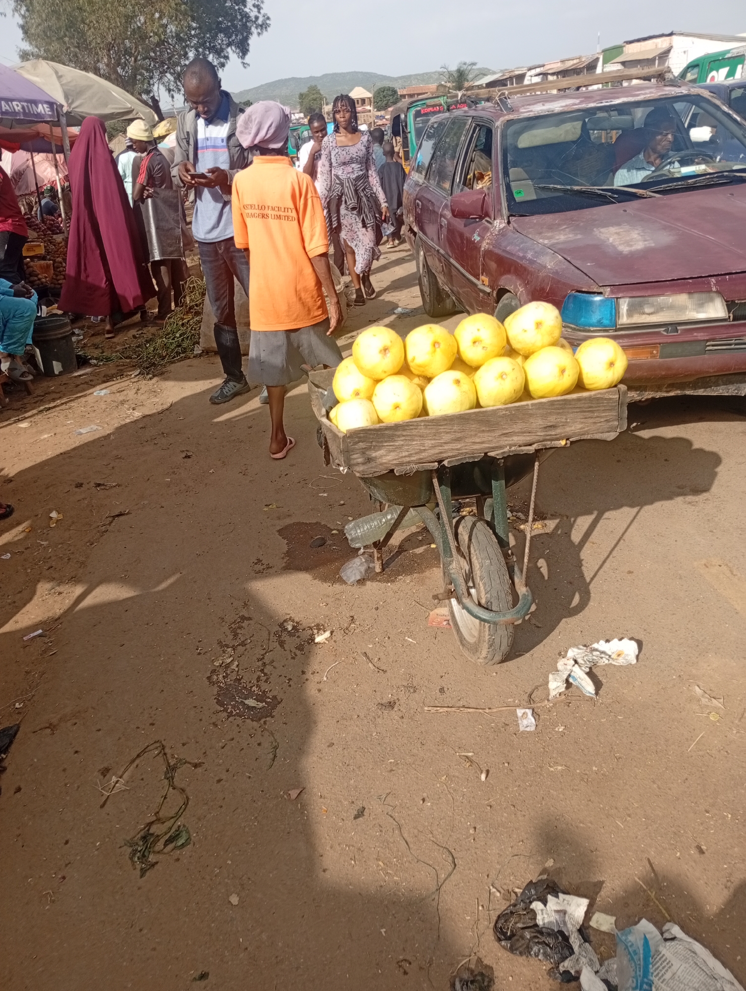 The image depicts an outdoor market scene in Rafin Par, Nigeria. A man wearing a blue and white striped shirt and dark pants is holding a smartphone, looking down at it. Beside him, a person in an orange t-shirt and grey shorts is standing, facing away from the camera. In the background, a maroon car is parked, with a man wearing glasses driving it. A wheelbarrow filled with yellow fruits is positioned in the foreground, casting a shadow on the dusty ground. Several other individuals and market stalls are visible in the background, suggesting a busy marketplace environment. The ground is composed of dirt and scattered debris.