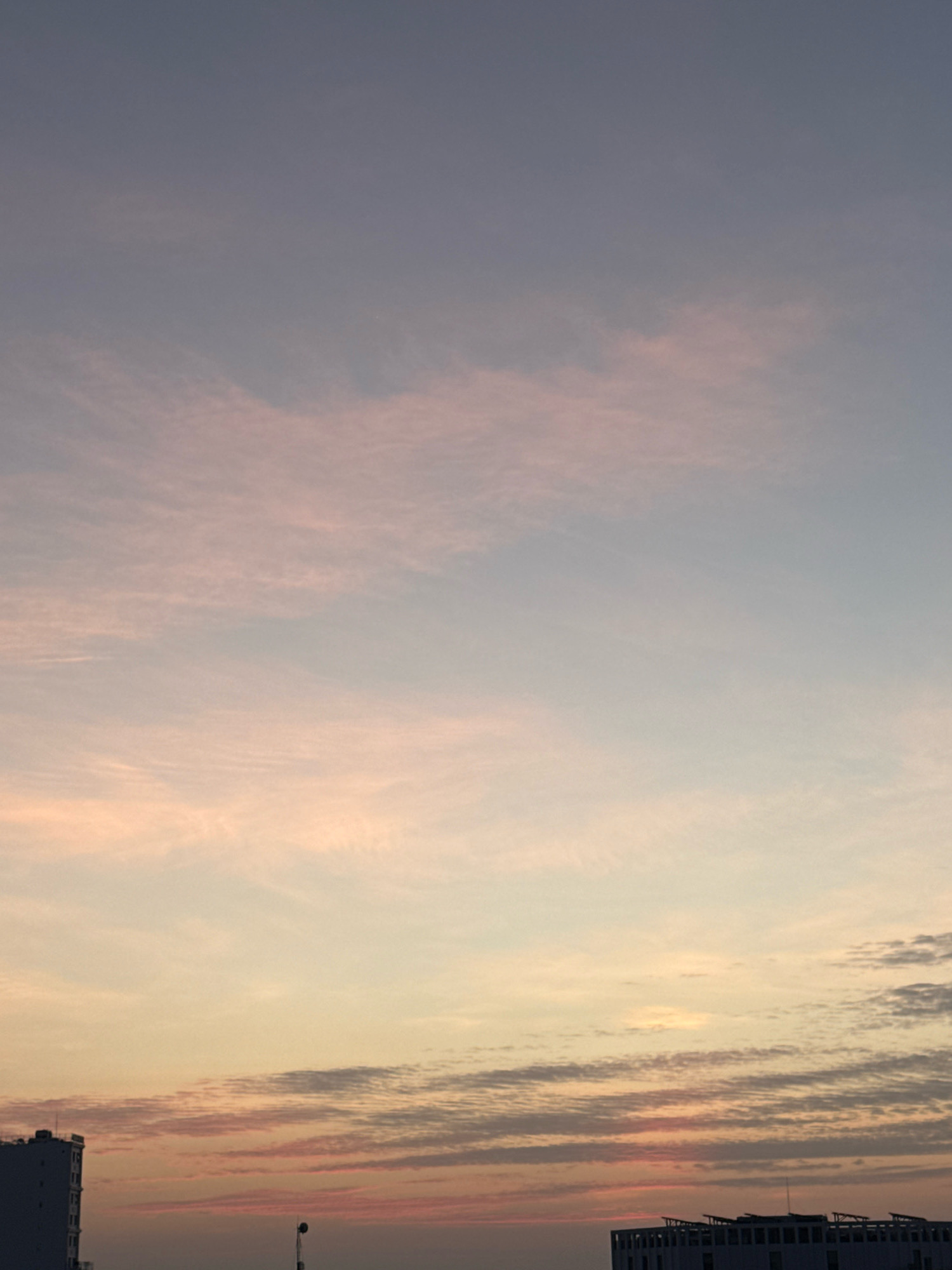 The image captures a sky at sunset or sunrise over the city of Nha Trang, Vietnam. The sky displays a gradient of colors, transitioning from a soft lavender at the top to pale yellow and pink hues closer to the horizon. wispy clouds are scattered across the sky, tinged with pink and orange. In the lower portion of the image, silhouetted against the colorful sky, are the tops of buildings. On the left, a tall, dark building with a flat roof is partially visible. To the right, another building with a more complex, multi-leveled roof structure is seen. The scene is devoid of any people or visible actions. The environment appears to be urban, with the buildings indicating a cityscape.