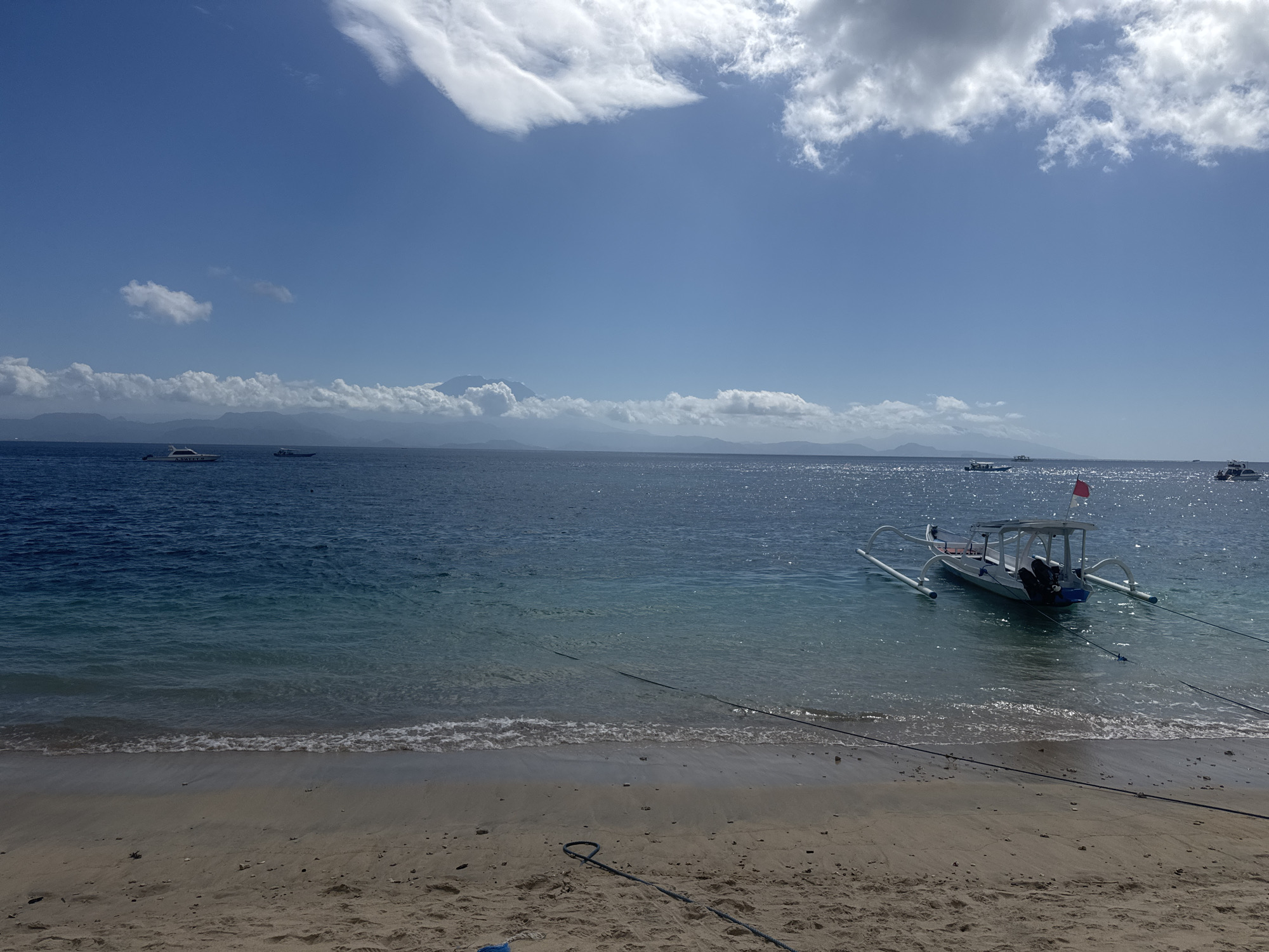 This is a medium-shot, horizontal, eye-level, outdoor photograph of a beach and the ocean. In the foreground, a sandy beach is visible, with a few small rocks scattered across it. A dark rope lies on the sand, forming a loop. In the middle ground, the turquoise water of the ocean gently laps at the shore. A traditional outrigger boat, painted white and blue with a canopy and two outboard motors, is moored in the water. It is attached to the shore by several ropes. In the background, the deep blue ocean extends to the horizon, where a chain of islands or a coastline is visible. Distant mountains can be seen on the horizon, with one prominent peak appearing to be a volcano, possibly Mount Agung. The sky is a bright blue, with scattered white clouds, some of which are wispy and stretched out. There are several other boats scattered across the water in the distance, including a white yacht-like vessel on the left. The overall scene depicts a tranquil tropical beach setting in Nusa Penida, Indonesia.