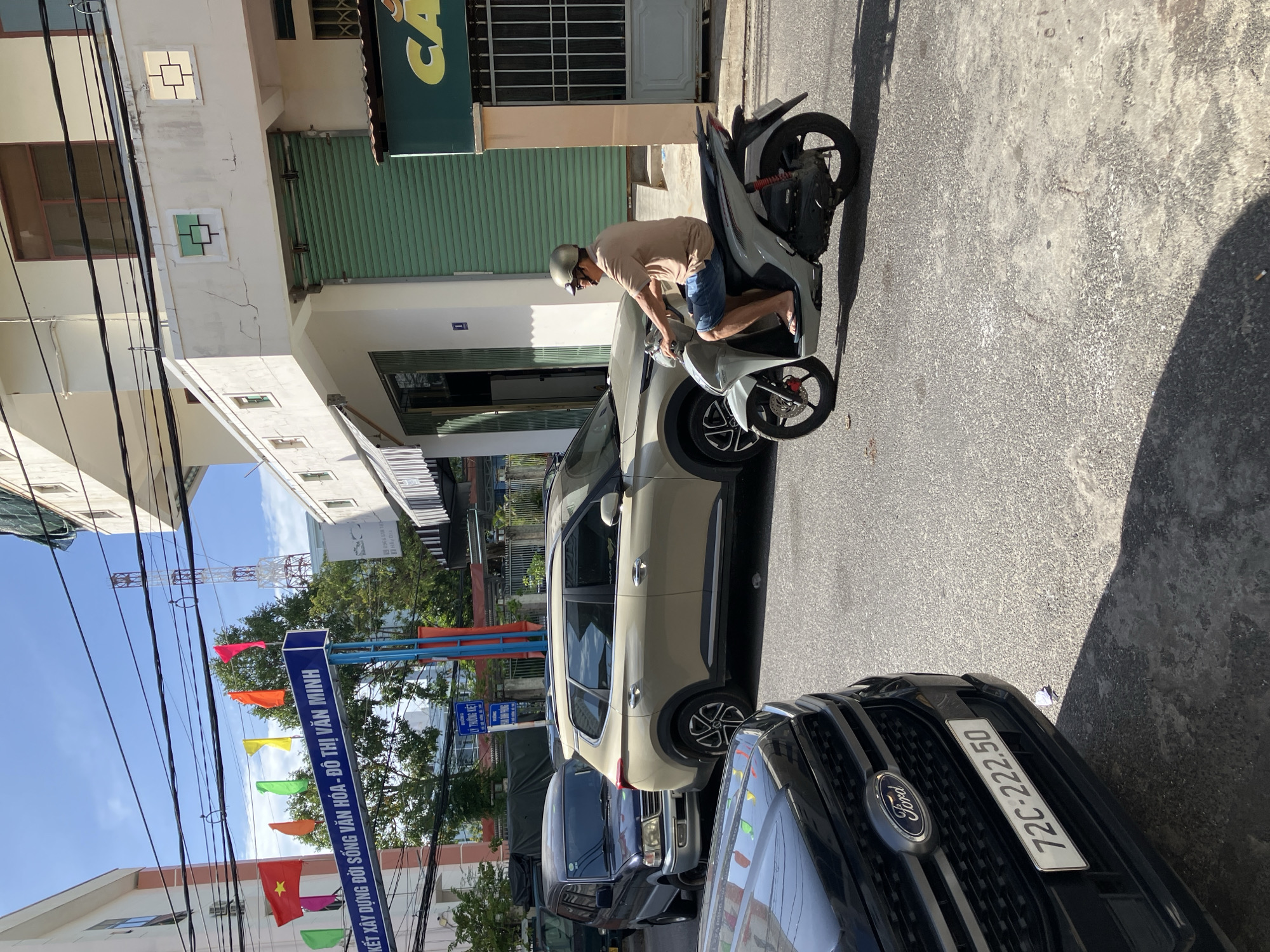 In a street scene in Nha Trang, Vietnam, a light-colored scooter is ridden by a man wearing a beige t-shirt, blue shorts, a helmet, and flip-flops. The man is looking forward as he rides. Behind him, a light beige SUV is parked or moving slowly. In the foreground, the front of a black Ford SUV is visible, displaying a license plate that reads "72C-222.50". The street is paved with asphalt. Buildings line the street, some with awnings and metal shutters. Colorful flags are displayed on poles above a blue sign that reads in Vietnamese "XÂY DỰNG ĐỜI SỐNG VĂN HÓA - ĐÔ THỊ VĂN MINH". The sky is blue with some clouds, and sunlight casts shadows on the ground. The scene appears to be captured during the daytime.