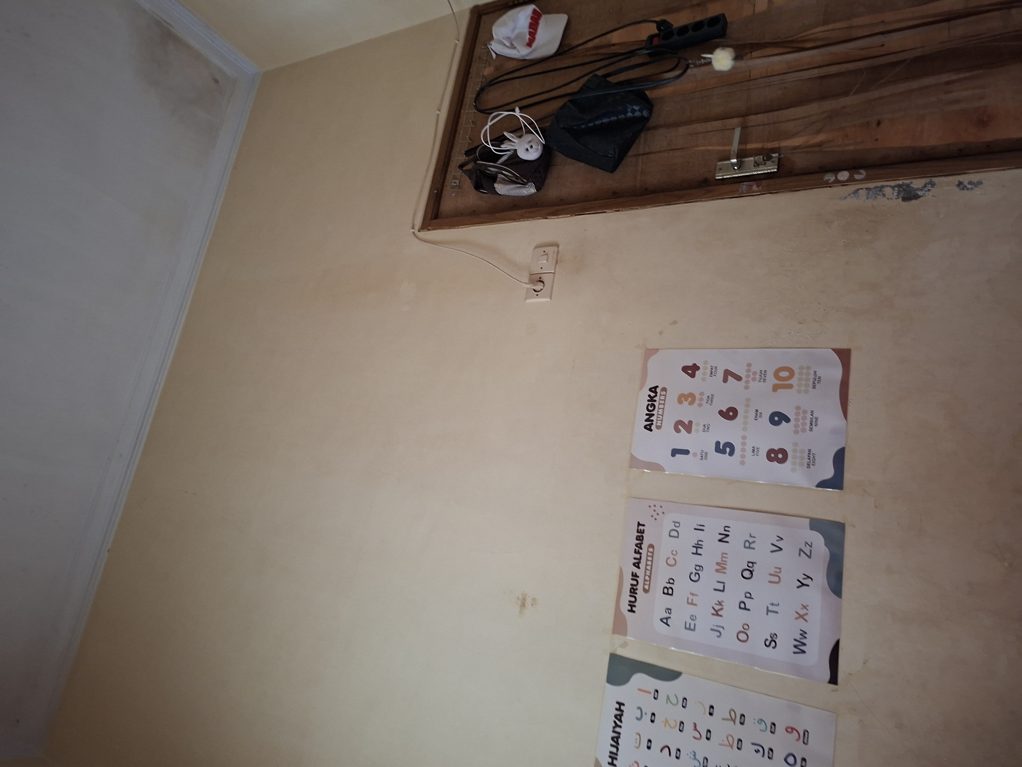 The image depicts an interior view of a room with cream-colored walls. On the left, three educational posters are affixed to the wall. The leftmost poster, titled "HIJAIYAH," displays Arabic letters. The middle poster, "HURUF ALFABET," shows the English alphabet from A to Z. The rightmost poster, "ANGKA," presents numbers 1 through 10, with their corresponding Indonesian names and dots.
To the right of the posters, a wooden door is visible, with several items hanging from hooks on its surface. These include two bags, one black and one with a striped pattern, a white cap with red lettering, and a white plush toy resembling a rabbit. A jump rope is also hanging, with its handles visible.
A light switch and an electrical outlet are mounted on the wall to the left of the door. The ceiling is visible in the upper portion of the image and appears to have some discoloration or water stains. The overall environment suggests a domestic or educational setting. The location is identified as Sekarpuro, Indonesia.