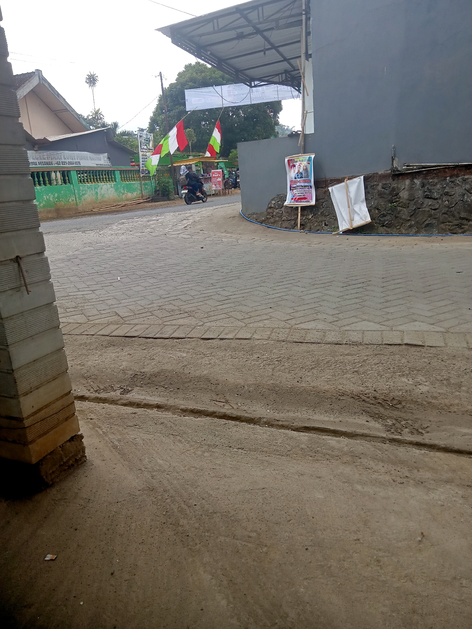 The image shows a street scene in Pule, Indonesia, with a paved road and a sidewalk. There are several objects and elements visible in the image.
On the left side of the image, there is a stack of concrete blocks, partially obscuring a green fence. The fence appears to enclose a property with some trees and buildings behind it. A sign on the fence reads, "SAJIAN IKAN LUAR LUHUR MUNGKAR RAU." Below this, a phone number, "+62 821-2664-9575," is visible.
In the middle of the road, a person is riding a motorcycle. The motorcycle is dark in color, and the rider is wearing a helmet and dark clothing. Two flags, one red and white and another green and white, are flying from a pole near the motorcycle.
On the right side of the image, a gray building with a stone foundation is visible. A banner with pictures of people is displayed on an easel in front of the building. Another white banner is also leaning against the stone foundation. A small sign that reads "HADI'S" is also visible.
The road is paved with interlocking concrete blocks, and there are tire tracks visible on the surface. The environment appears to be a relatively busy street, with buildings and trees lining the road. The sky is overcast, suggesting it might be a cloudy day.