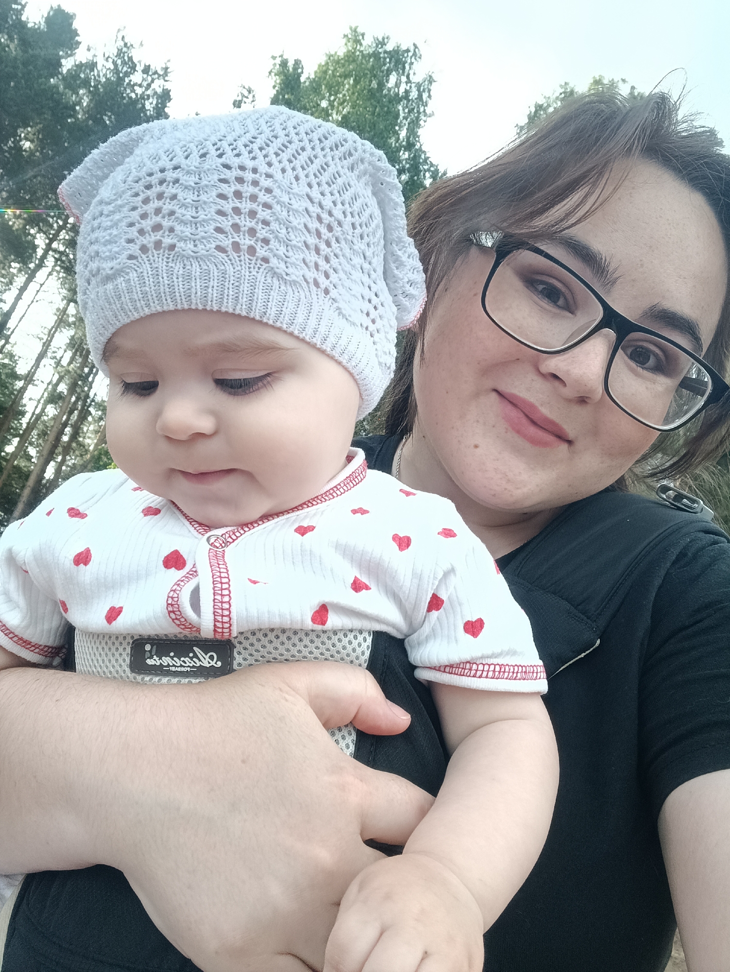 A young woman with dark brown hair and glasses holds a baby wearing a white knit hat and a white shirt with red hearts. They are outdoors, with trees in the background. The woman is wearing a black top. The baby is in a carrier. The scene appears to be a park or wooded area.
