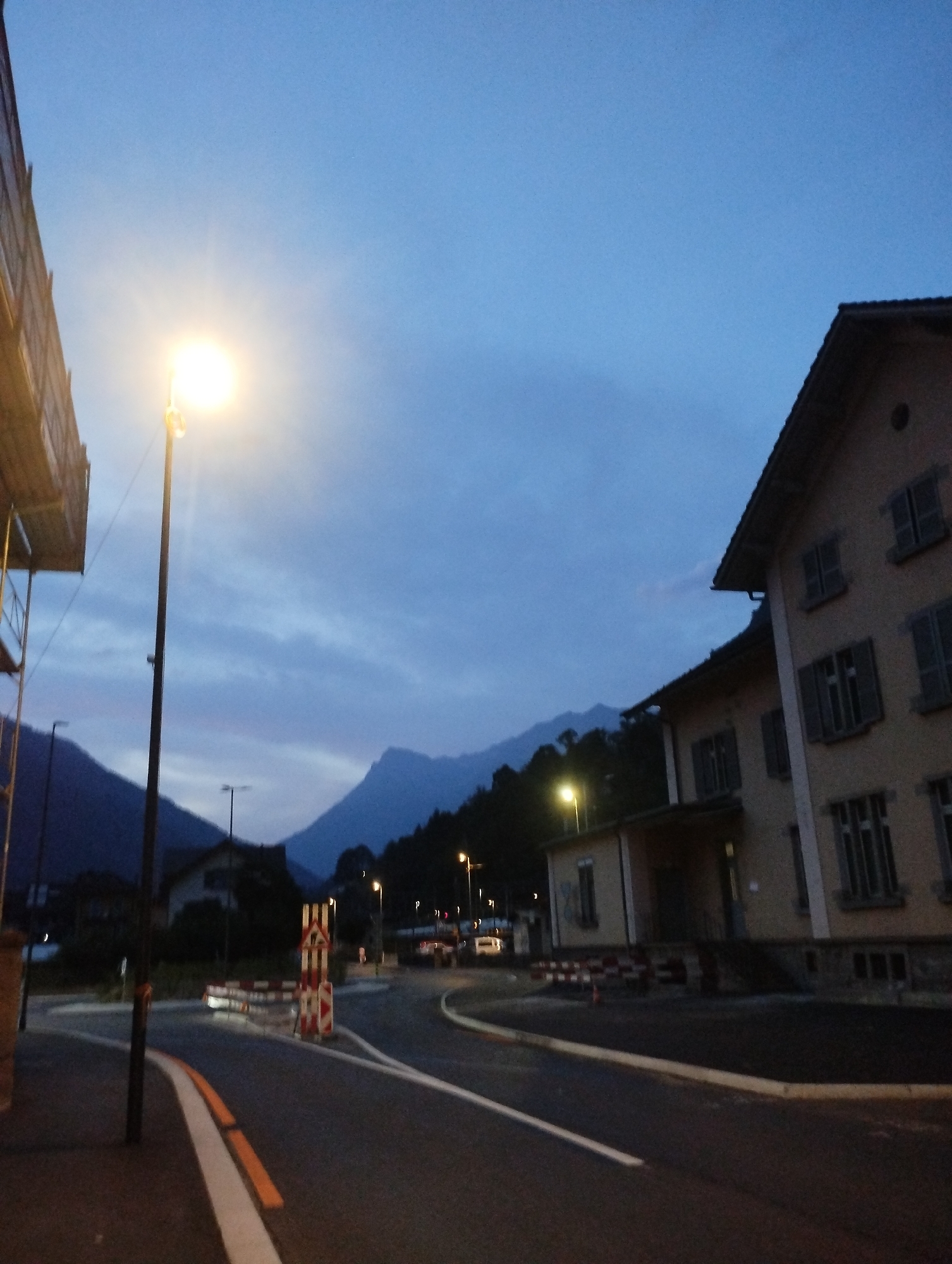 An eye-level, full shot captures an evening scene on a street in an unknown town, possibly at dusk due to the twilight sky and illuminated streetlights. The road is paved and curves to the right, with white and orange markings indicating lanes and edges. Construction barriers, a mix of red and white striped traffic signs and barriers, are present on the side of the road.
On the right side of the street, a two-story building with a light-colored facade stands prominently. The building has a dark, sloped roof and multiple windows with dark shutters. The windows appear to be symmetrically placed on each floor.
To the left of the road, a tall, dark lamppost with a brightly lit lamp illuminates the immediate area. Beyond the lamppost, scaffolding is visible attached to an unseen structure, suggesting ongoing construction. In the background, distant mountains are silhouetted against the dusky sky. The overall atmosphere is quiet and peaceful, with the only significant light sources being the streetlights and the building's interior lights, which are not clearly visible. No people are present in the scene.