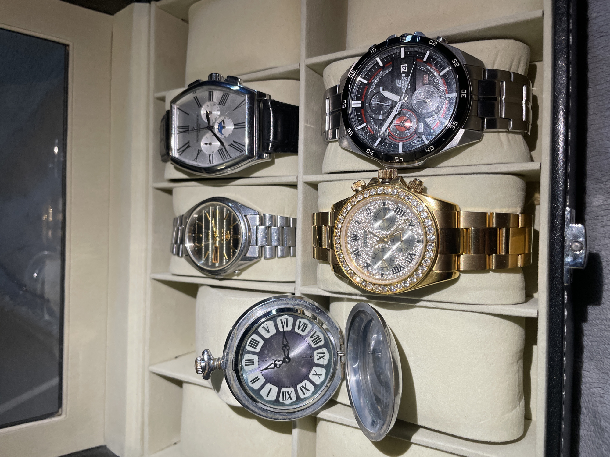 The image shows a collection of six watches displayed inside a watch box with a window on the left. The watch box has individual compartments for each watch, with padded cushions to hold them in place.
From top left to bottom:
1. A rectangular silver-colored watch with a black leather strap.
2. A large, black-faced chronograph with a stainless steel bracelet. The watch face features red accents and the word "EDIFICE".
3. A silver-colored watch with an intricate, partly see-through dial and a silver bracelet.
4. A gold-colored watch with a diamond-studded bezel and dial, with a gold-colored bracelet.
5. A silver pocket watch with a gray face, Roman numerals, and its front lid open.