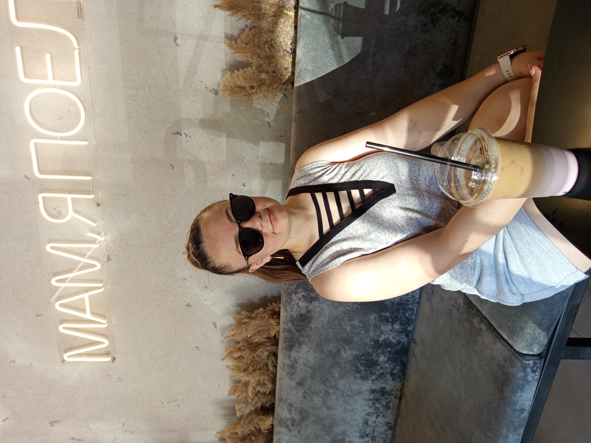 A woman sits on a grey couch in front of a concrete wall with a neon sign that reads "МАМ, Я ПОЕЛ" which translates to "Mom, I ate". She is wearing sunglasses and a grey tank top with black straps across the chest. She is holding a clear plastic cup with a straw containing an iced coffee. Dried plants are visible behind her in planters. The lighting suggests it is daytime, with shadows cast on the wall and the woman.
