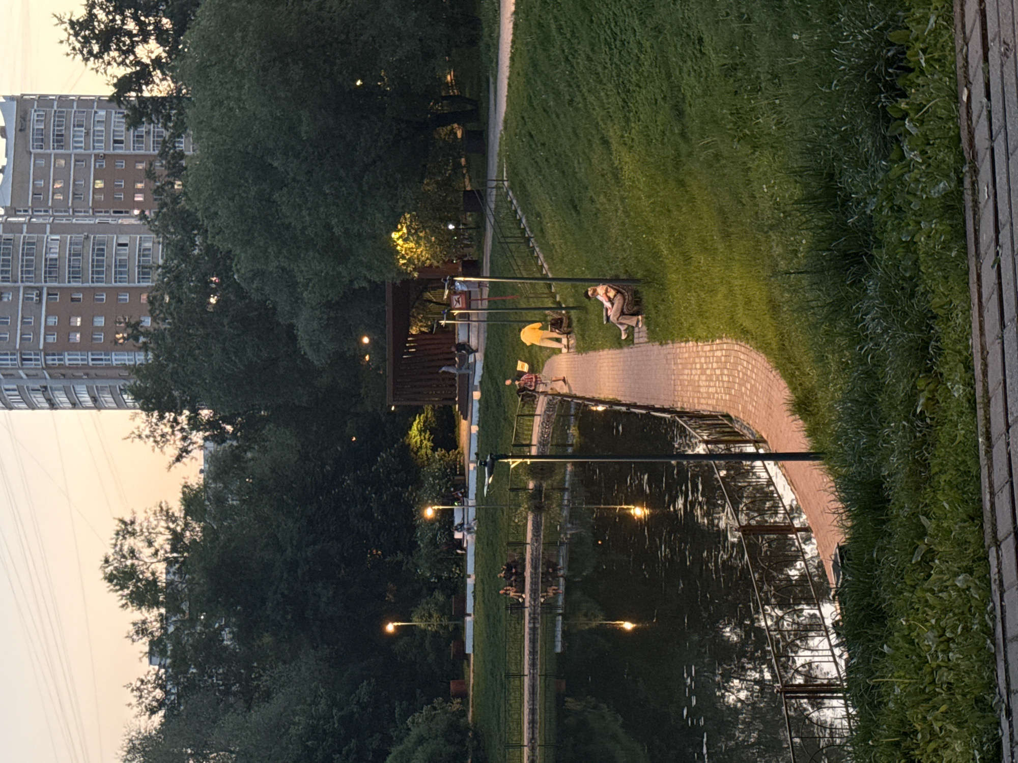 The image depicts a park setting, potentially in Moscow, Russia, given the architectural style of the building visible. A pond or lake reflects the surrounding trees and lampposts, creating a symmetrical visual effect. A brick pathway runs along the water's edge, where individuals are seen engaging in leisure activities, such as sitting on benches and strolling. The park appears to be illuminated by both natural light and the warm glow of lampposts, suggesting it is either dusk or dawn. A multi-story residential building is situated in the background, partially obscured by trees, hinting at the park's location within an urban environment. The greenery consists of well-maintained grass and trees, which contribute to a tranquil atmosphere.