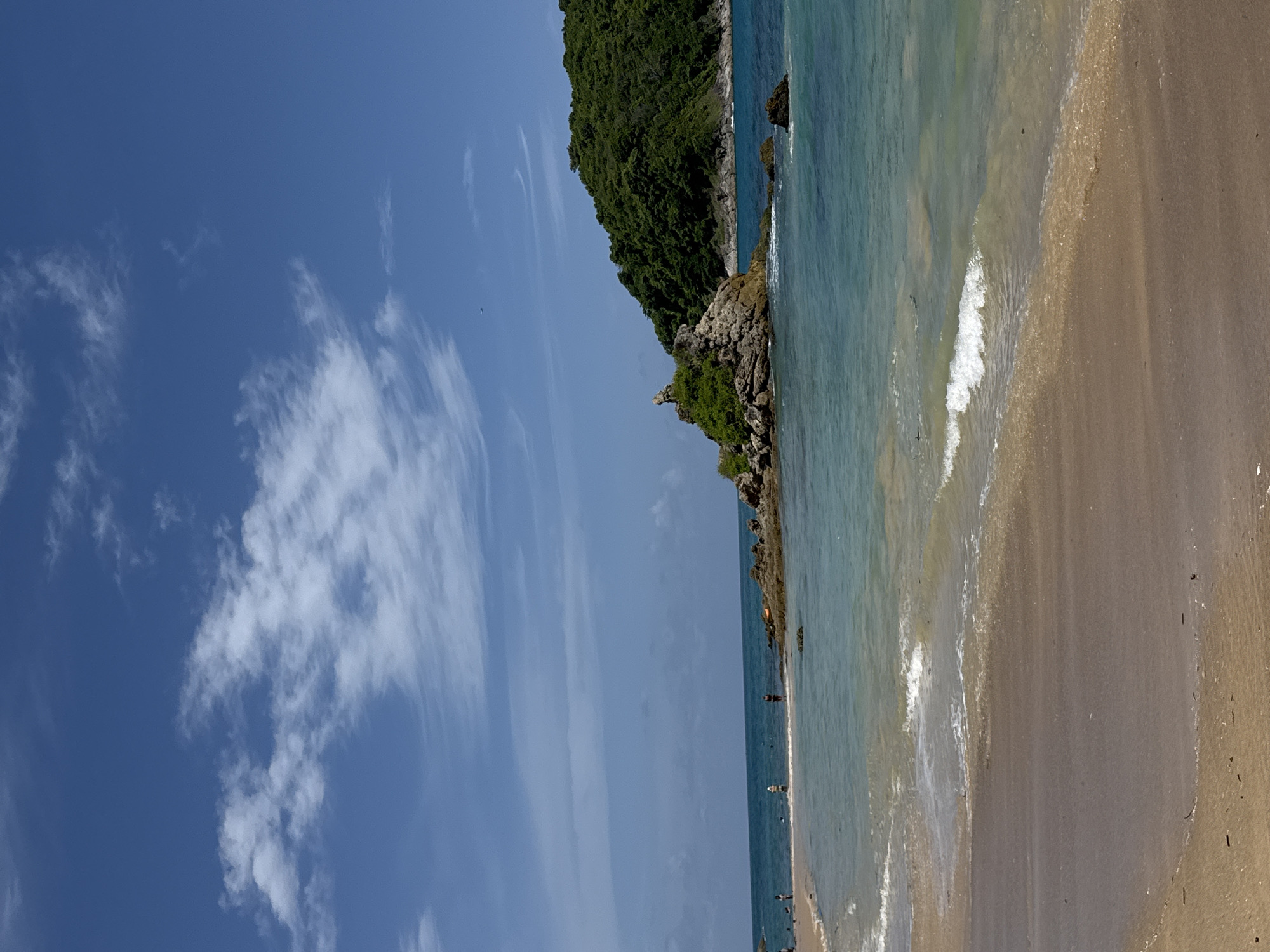 The image depicts a bright, sunny day at Rawai Beach, Thailand. The scene is dominated by a clear blue sky with scattered white clouds. The foreground is a sandy beach, with gentle waves lapping at the shore. In the midground, the turquoise water stretches out, with a few small rocks and a larger rocky outcrop covered in vegetation situated in the sea. Further in the distance, a lush green, tree-covered island rises from the water. Several people are visible along the shoreline, some wading in the shallow water and others standing on the beach. The overall impression is of a tranquil tropical beach scene.