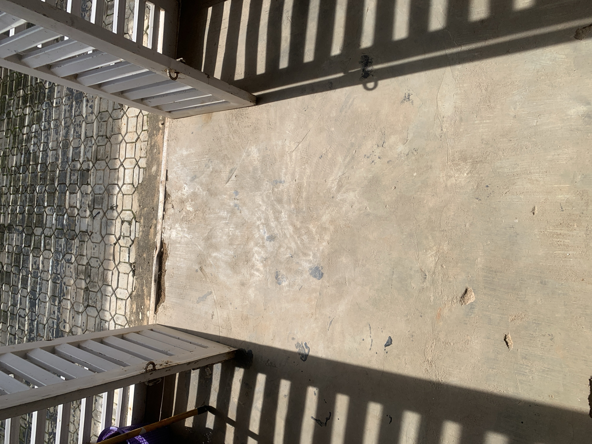 An outdoor scene shows two white louvered gates, one on the left and one on the right, opening inward towards the viewer. The gates are casting long, parallel shadows across a concrete floor. Beyond the gates, a patterned stone walkway is visible under bright sunlight. The concrete floor appears worn and uneven, with several dark smudges and areas of discoloration. A purple laundry basket and a wooden-handled tool are partially visible to the left of the left gate. The scene is devoid of any people.