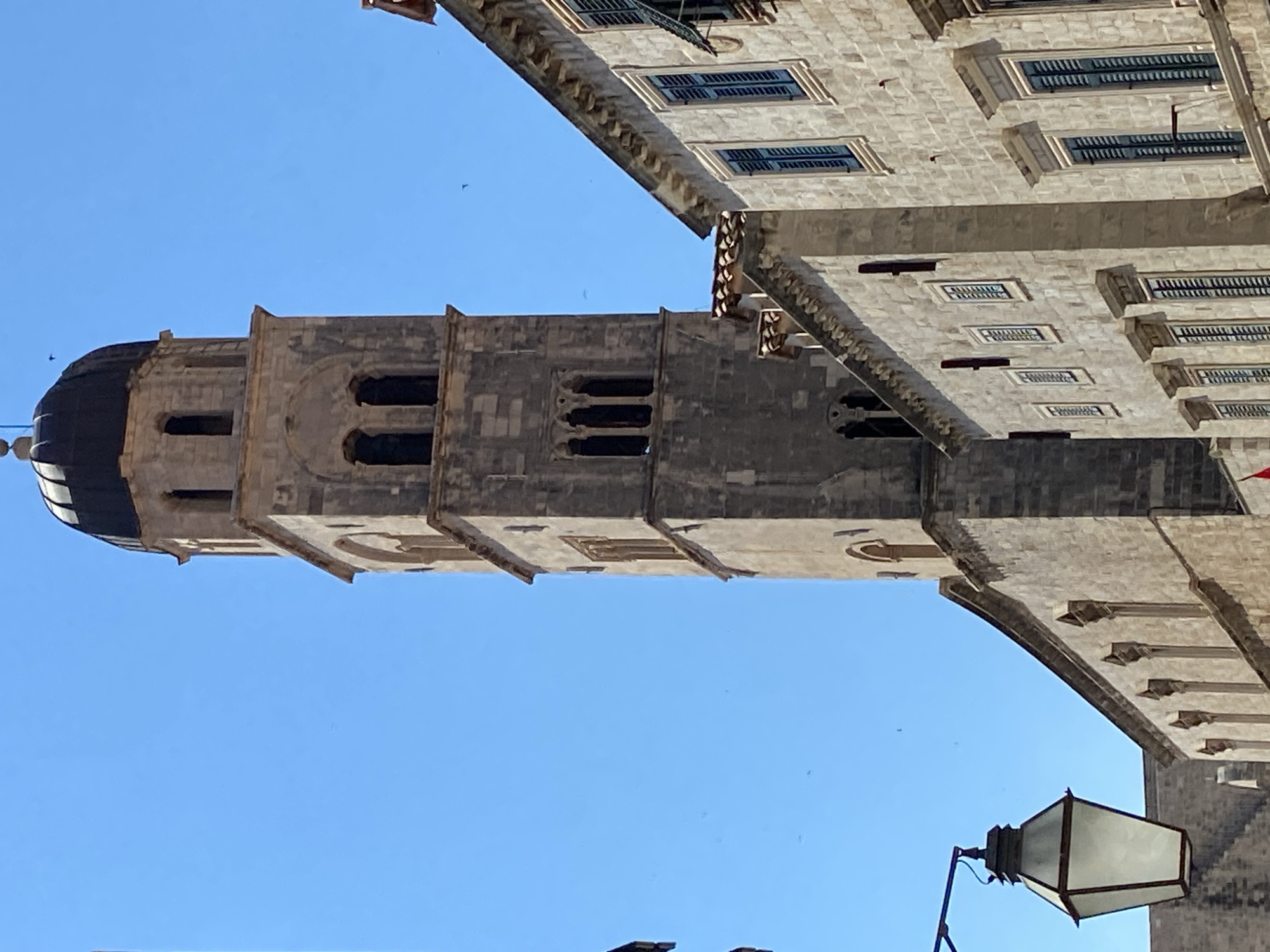 The image shows a stone bell tower with a dark dome against a clear blue sky, situated in Dubrovnik, Croatia. The tower is partially obscured by a building made of similar stone with multiple windows, some featuring dark shutters. A street lamp with a white lantern is visible in the lower left corner, hanging from a black metal arm. The architecture suggests a historic European city.