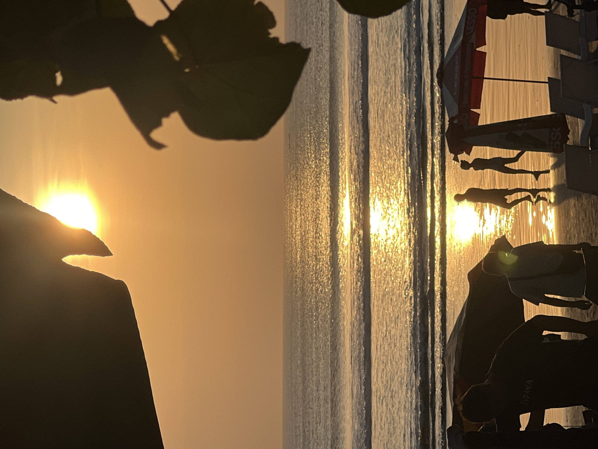 This is a photograph of Kuta Beach in Kuta, Indonesia, during sunset. The sun is low in the sky, casting a warm, golden glow across the scene. The ocean is calm with gentle waves, reflecting the sunlight. In the foreground, silhouetted figures of people are visible on the beach. Some appear to be walking, while others are standing. There are also beach chairs and an umbrella, suggesting a recreational area. Foliage from a tree partially obscures the top of the frame, framing the sun. The overall atmosphere is serene and tranquil, capturing the essence of a tropical sunset.