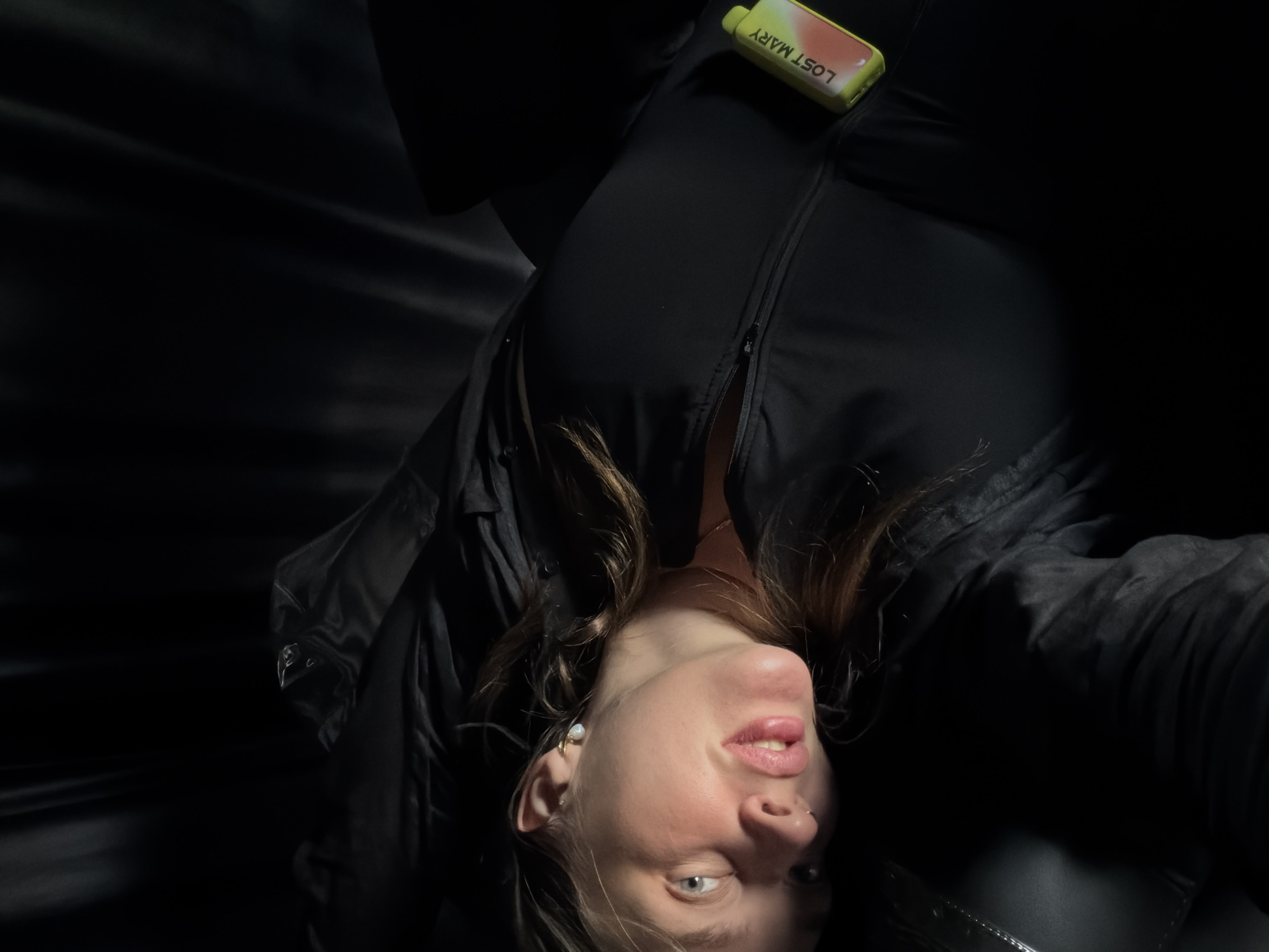 A woman is lying on her back on a dark surface, possibly a couch or bed, with her head turned towards the camera. She has light brown hair, light blue eyes, and full lips. She is wearing a black top and a jacket or coat, also black. A vape device with the label "LOST MARY" is visible tucked into the front of her clothing. The background is dark and indistinct, with fabric folds visible, suggesting an indoor setting. The lighting is dim, with a spotlight illuminating her face and upper body. There are no visible indications of the specific location in Укромновское сельское поселение, Ukraine.