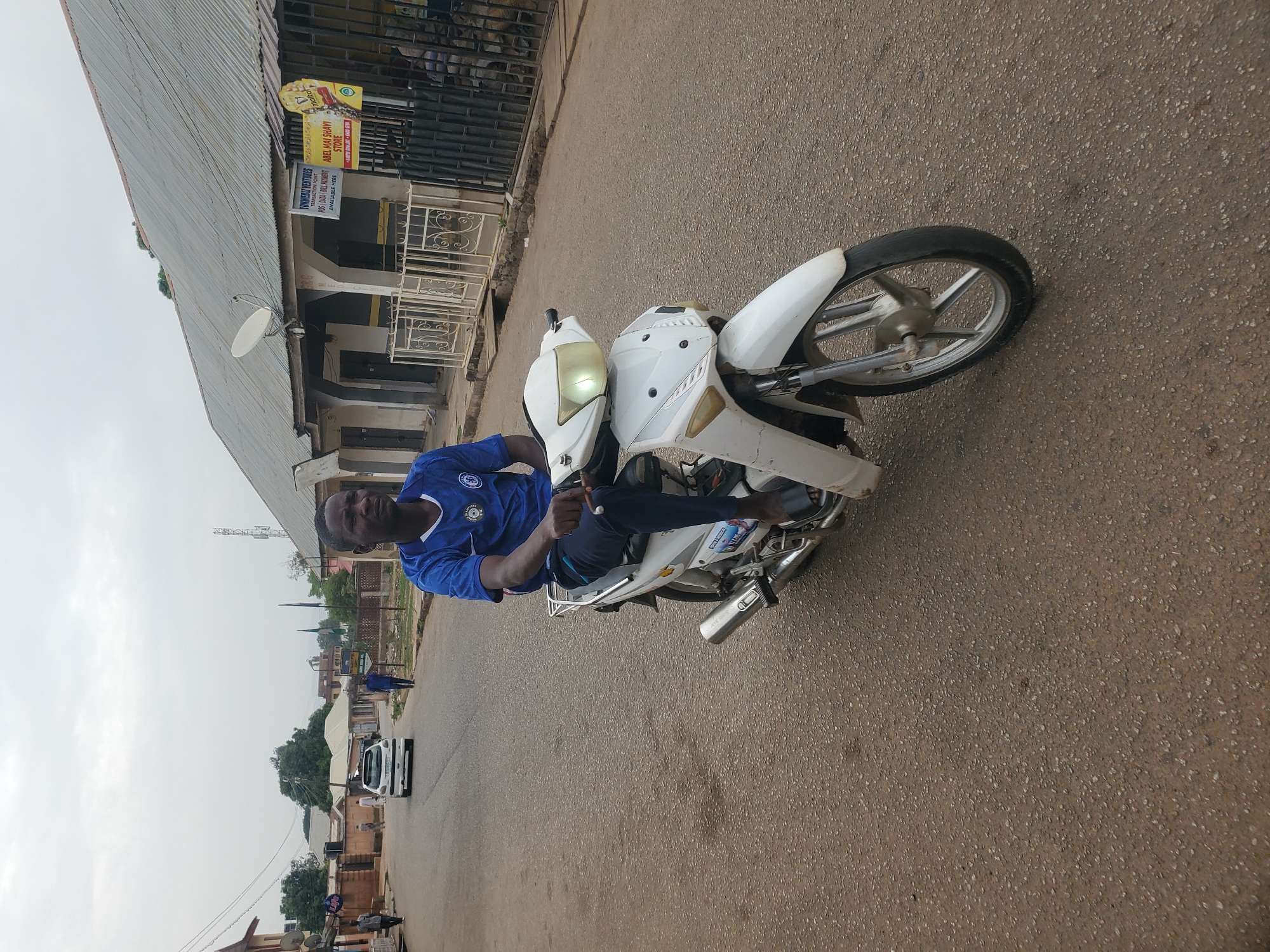 The image shows a man riding a white motorcycle on a paved road. He is wearing a blue shirt with a Chelsea FC emblem. The background includes buildings with corrugated metal roofs and a sign reading "FONKIEBIZ VENTURES TRANSACTION POINT POS DATA BILL PAYMENT AVAILABLE HERE". A vehicle can be seen driving on the road behind the man. The location is indicated by the sign as being in the city of Jos, Nigeria.