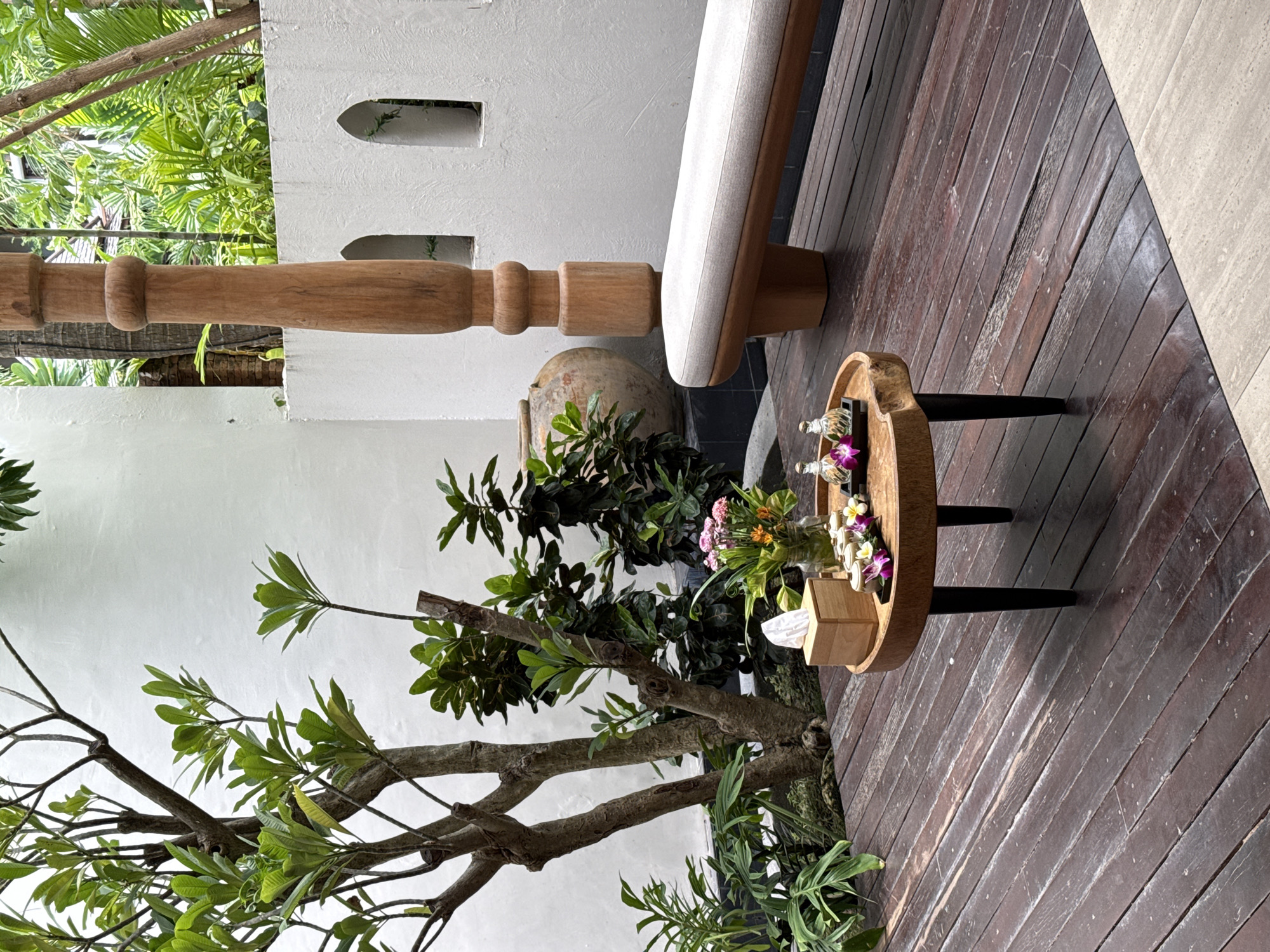 The image depicts an outdoor serene setting, likely a spa or resort area in Ko Samui District, Thailand. In the foreground, a circular wooden table with black legs holds a wooden tissue box, a small vase of flowers, and two glass bottles containing a clear liquid, possibly massage oils. The table is placed on a weathered wooden deck. To the right of the table, a light-colored bench with wooden legs is visible. Behind the bench, a tall, intricately carved wooden pillar stands. The background features a white textured wall with arched openings, beyond which lush green foliage is visible. A large, textured pottery vessel is placed against the wall. The overall environment suggests a tranquil and natural ambiance.