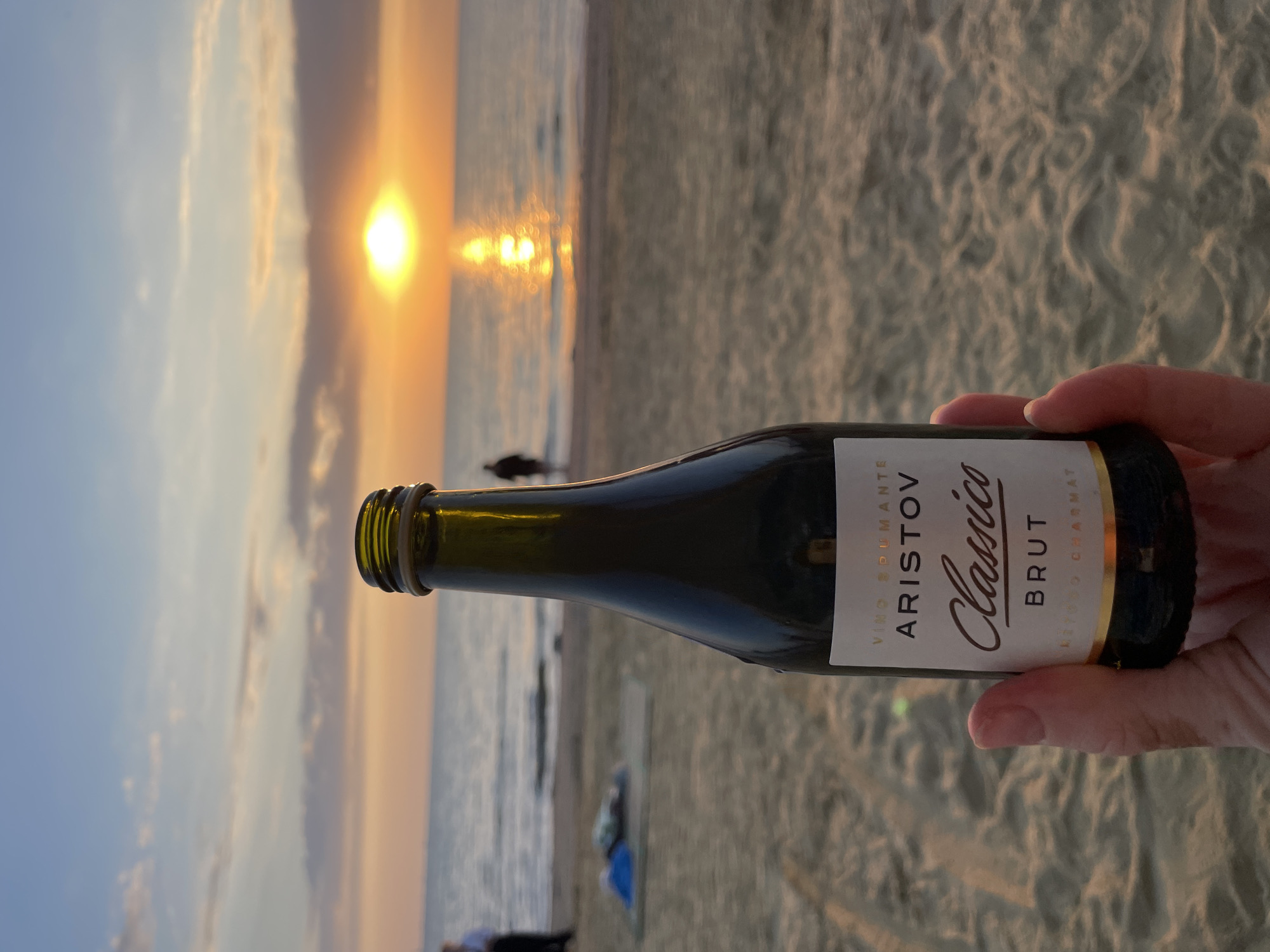 The image shows a person's hand holding a bottle of Aristov Classico Brut wine on a sandy beach in Anapa, Russia. The sun is setting over the Black Sea, casting a warm glow on the water and sand. The sky is a mix of orange, pink, and blue hues, with some scattered clouds. In the background, there are a few people walking along the shoreline and some beach equipment. The bottle is dark green glass with a white label that reads "VINO SPUMANTE ARISTOV Classico BRUT". The label is partially obscured by the hand holding the bottle. The scene evokes a sense of relaxation and enjoyment of a sunset by the sea.
