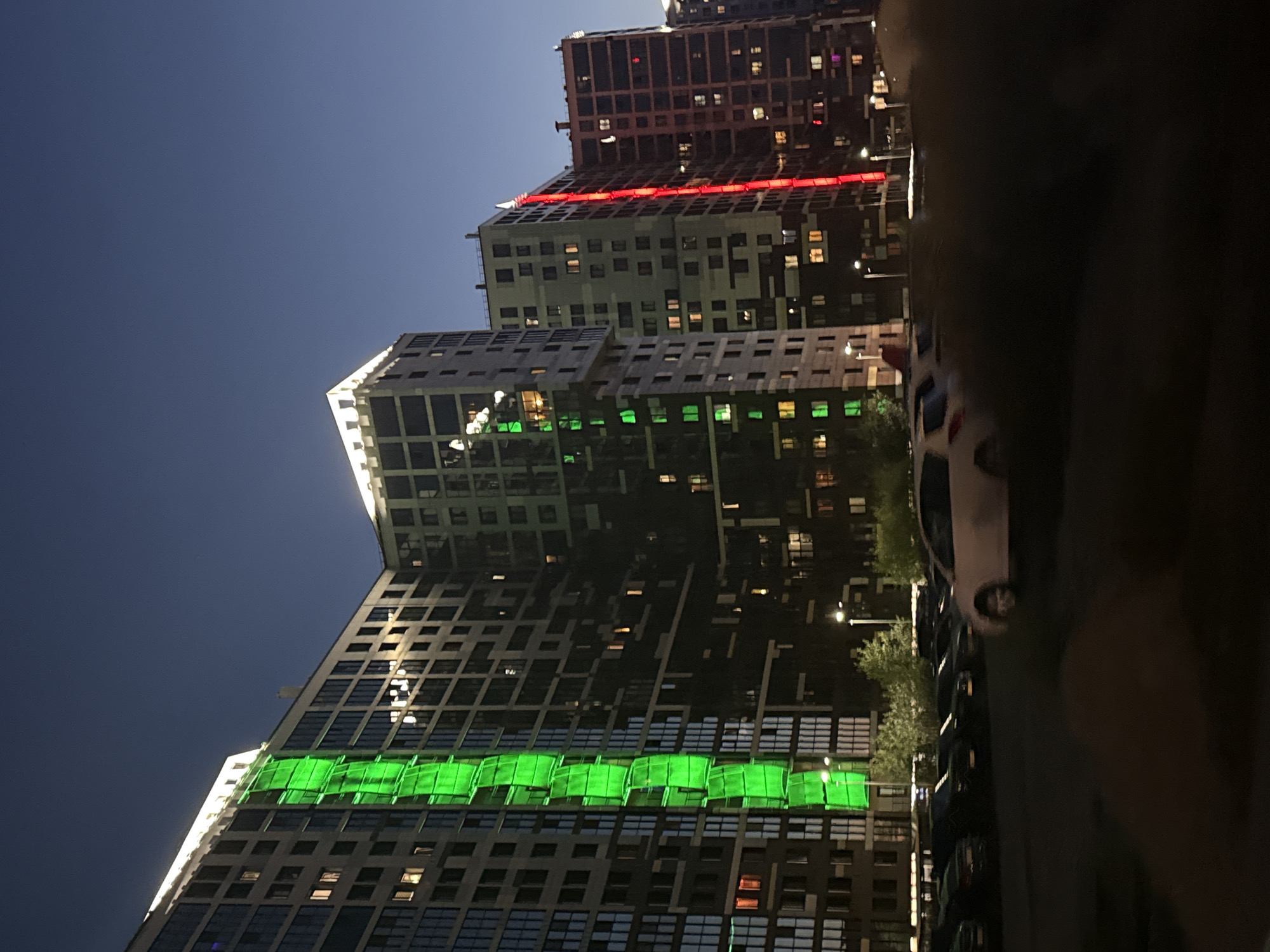 The image depicts a nighttime scene in Saint Petersburg, Russia, focusing on a row of modern, multi-story residential buildings. The buildings are illuminated, with prominent vertical bands of bright green light on the leftmost building and red light on a building further to the right. The sky is a deep, clear blue, characteristic of twilight or night. In the foreground, a parking lot is visible, containing several cars, some of which are white. The lower portion of the image is dark and somewhat blurred, obscuring finer details of the ground and immediate surroundings, but suggesting a pedestrian area or shrubbery. The overall impression is one of urban development with contemporary architectural features, including decorative lighting.