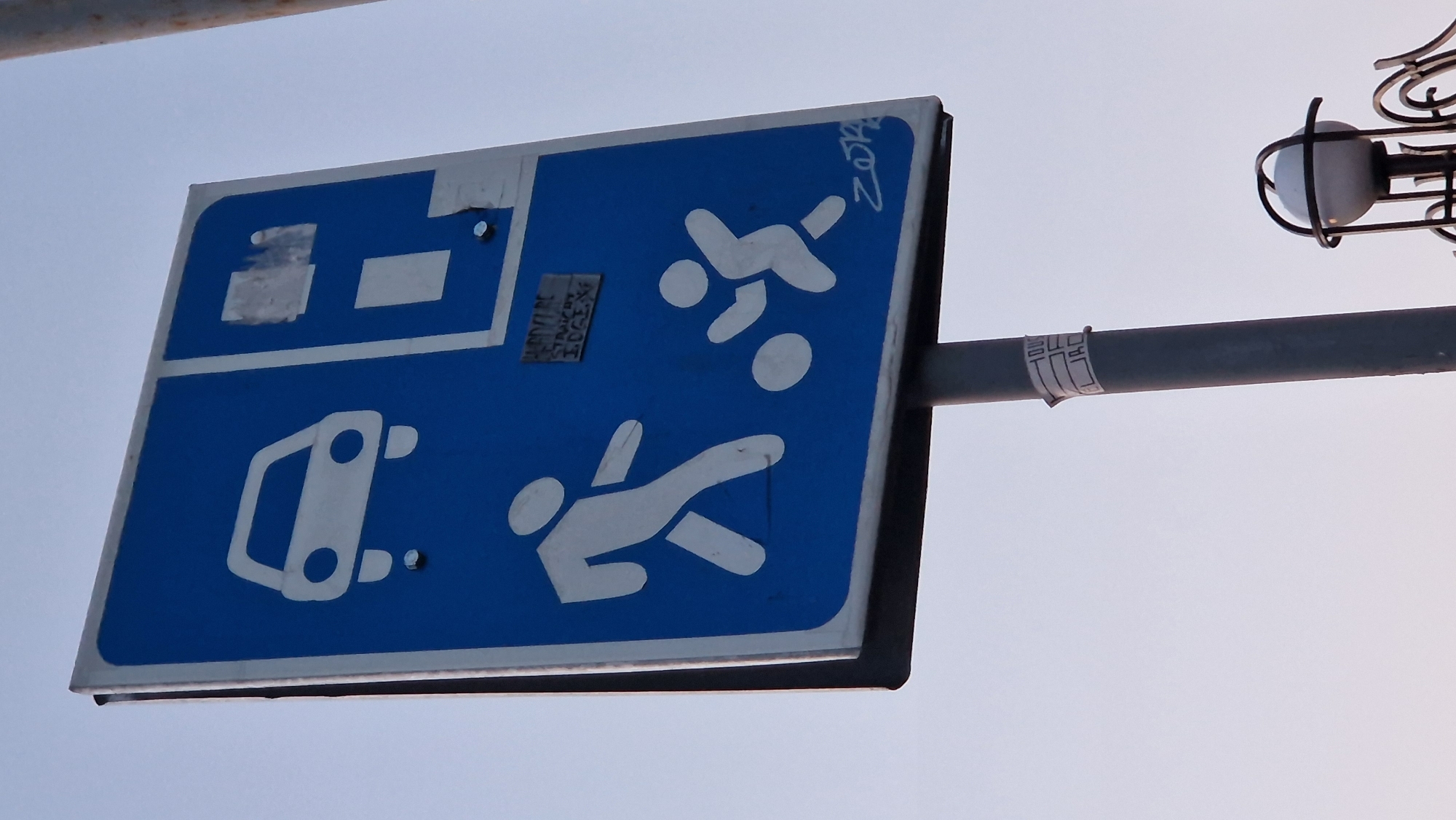A blue traffic sign with white symbols is mounted on a gray pole against a pale sky. The sign depicts a white outline of a car at the top left, and below it, two white figures are drawn. The figure on the left appears to be an adult walking, while the figure on the right is a smaller child playing with a ball. In the upper right section of the sign, there are two rectangular white boxes, partially obscured by what appears to be tape. There are also some handwritten markings in black on the sign, including a small tag with text and a graffiti tag at the bottom right. A sticker with black text is wrapped around the pole below the sign. A street light fixture is visible at the bottom right of the image.