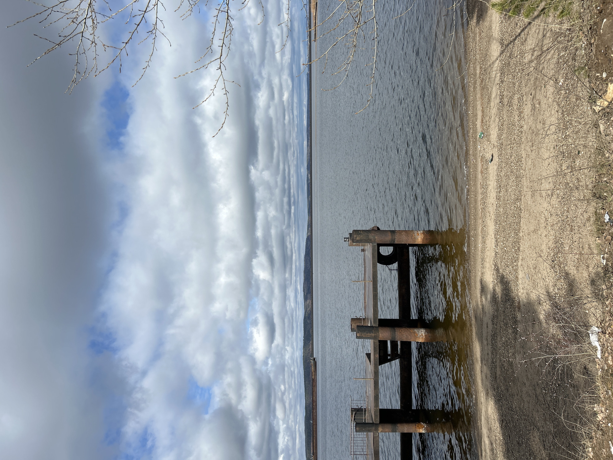This landscape photograph, taken in Chaikovsky, Russia, features a river or lake scene under a cloudy sky. The foreground shows a rusty, dilapidated pier structure made of metal pipes extending into the water. A beach stretches along the right side of the frame, blending from sand to dirt with sparse vegetation. A strip of land is seen in the mid-ground between the water and the horizon. The sky is dominated by a pattern of grey and white clouds, with a hint of blue peeking through. Bare tree branches are visible in the top left corner of the frame. The overall impression is a mix of industrial decay and natural beauty in a temperate climate.