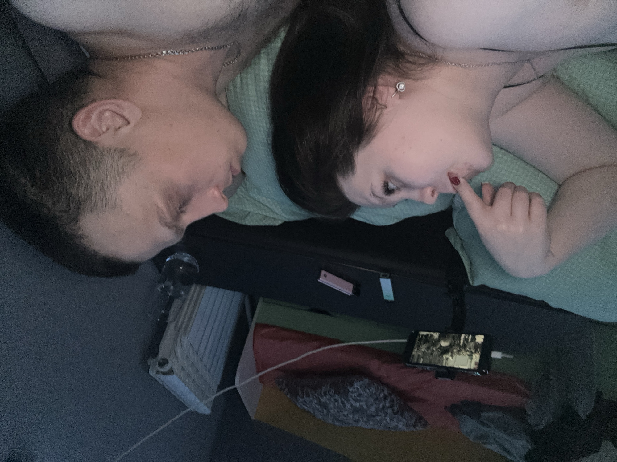 A man and a woman are lying next to each other on a bed. They are both facing each other. A smartphone displaying a video is charging next to them on the bed. A radiator is in the background. The location is Chaikovsky, Russia.