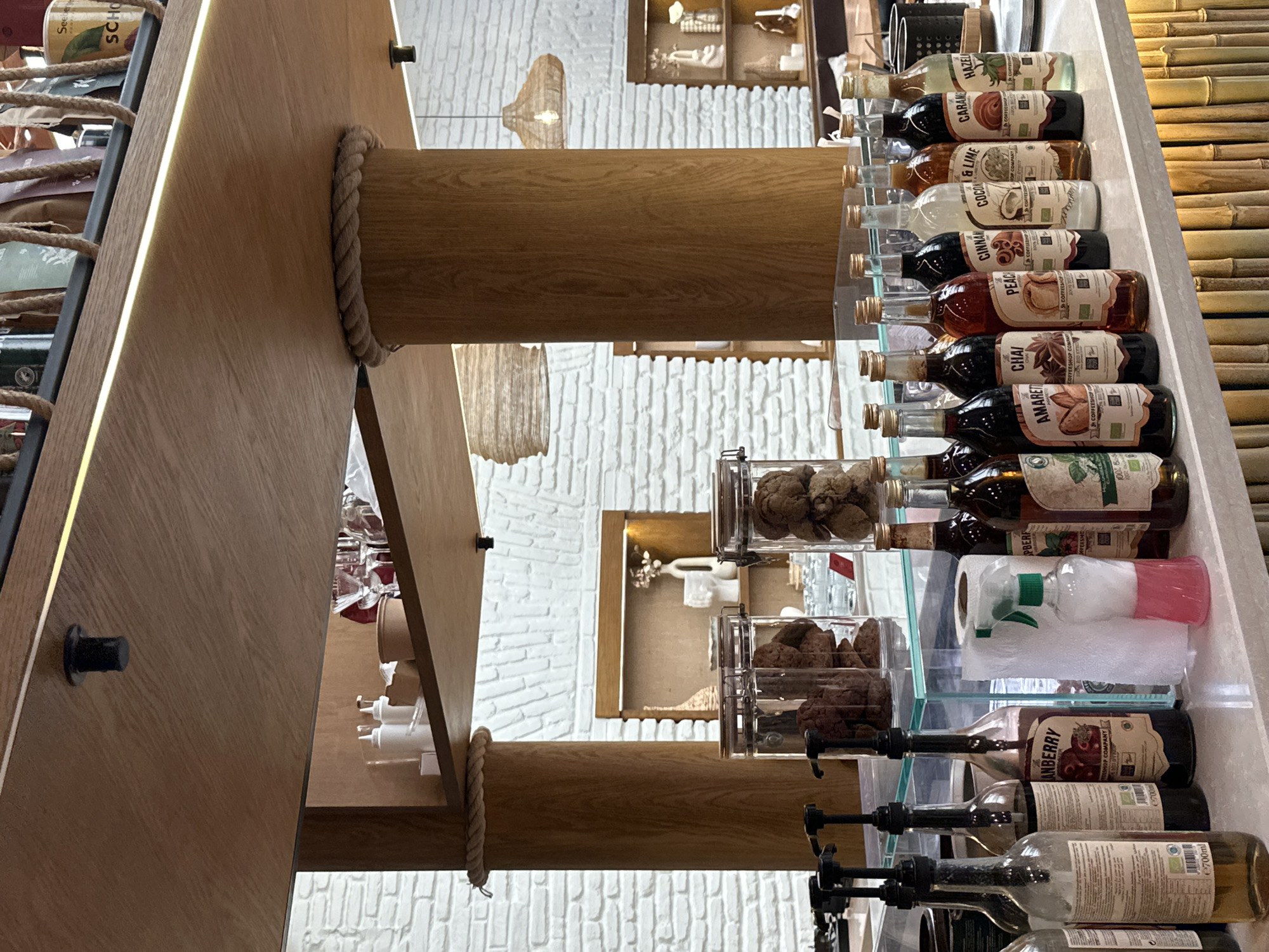 This is an indoor shot of a cafe or coffee shop, most likely in Tbilisi, Georgia. The camera angle is slightly upward.
The dominant features are a wooden support structure with thick rope wrappings and a counter lined with glass containers filled with cookies and rows of labeled bottles, potentially syrups or flavorings for drinks. Visible bottle labels read "HAZEL", "CARAME," "COCOA & LIME", "CINNA", "PEAC", "CHAI," "AMARET", and "ANBERRY," along with the text "COFFEESHOP COMPANY."
There is a cleaning spray bottle near the cookies and empty bottles with dispensers on the lower shelves. The wall behind is white brick with decorative shelves. In the far upper left corner, partially visible are the words "Seeber" and "SCHO" on a paper container. A bamboo wall is seen on the right.