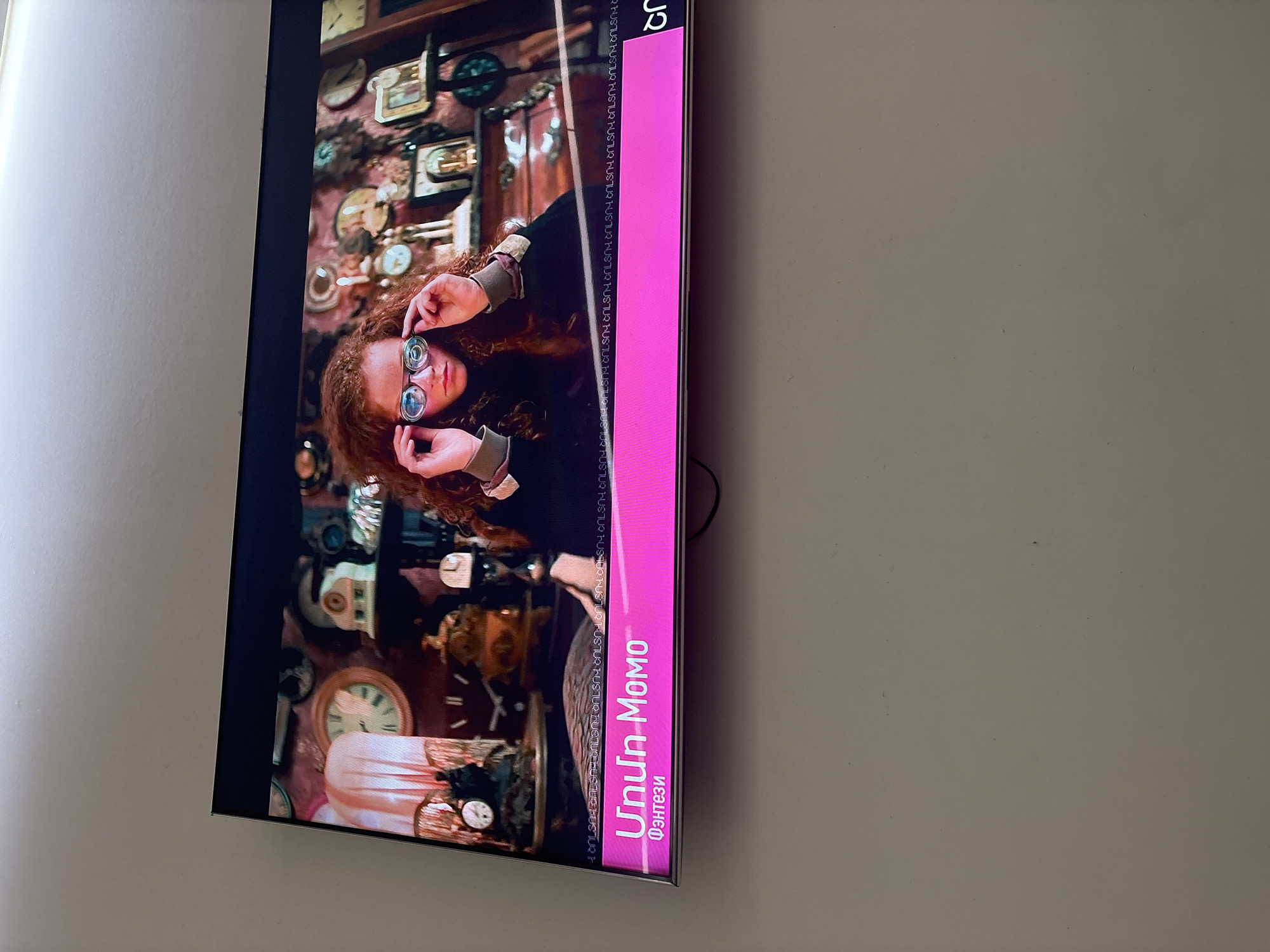 A television screen, tilted and viewed from an angle, is the central focus, displaying a scene from a movie or TV show. The screen shows a young person with curly red hair and glasses looking forward, holding another pair of round-lensed spectacles. The background of the scene is filled with numerous clocks, gears, and various timepieces, suggesting an antique shop, workshop, or a fantastical setting.
A prominent vertical pink bar occupies the right side of the TV screen, containing textual information. The main title "Unun MOMO" is displayed in large white letters, with "Фэнтези" (Russian for "Fantasy") written below it in smaller white text, indicating the genre. The letters "JQ" are visible in the top right corner of this pink bar. A stylized, repetitive pattern of text-like characters borders the video content on the left side of the pink bar, functioning as a decorative element.
The TV is mounted against a plain, light-colored wall, which is visible to the left and slightly below the screen. A dark cable can be seen behind the TV. The image is captured indoors.