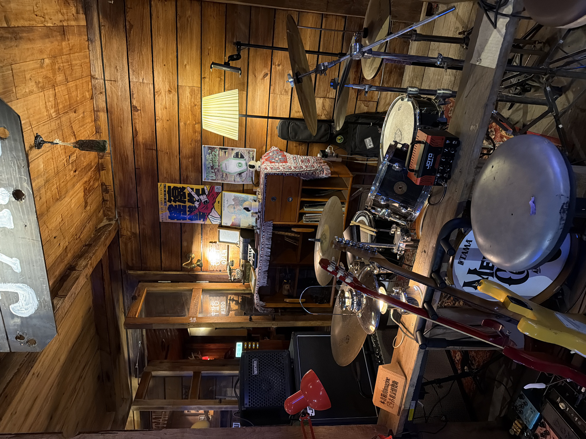 The image shows a cluttered music room, likely in a personal residence, given the wooden walls and beamed ceiling. The location may be Chongqing High-tech Zone, China, due to the Chinese text on a wooden block.
Musical equipment is abundant: drums, cymbals, guitars, and amplifiers are prominently visible. A "Warwick" amplifier and "JOYO" brand equipment are among the brands seen. A "TAMA" brand logo is visible on the drum kit.
Other objects include posters on the wall, a lamp, a table covered with a patterned cloth, and a red desk lamp near the foreground. Light fixtures are also visible near the ceiling. The arrangement of items suggests a small, possibly confined space, with a mix of musical equipment and everyday objects.