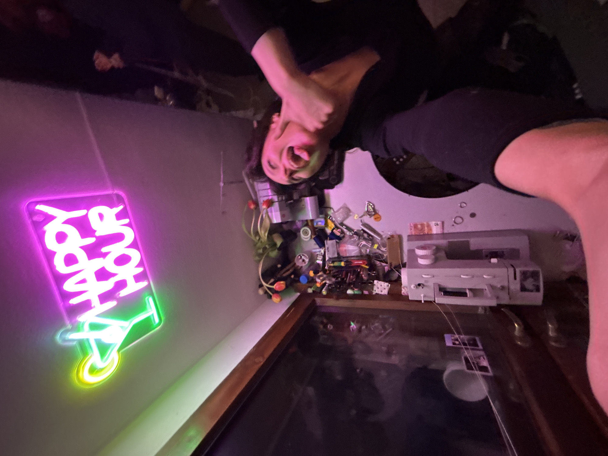 A person is upside down with their head near a table. The person is light-skinned and wearing dark clothing. They have their hands around their neck and their mouth is open, possibly yelling. On the table is a collection of objects including a gray electronic device, a plant, various small items, and a neon sign reading "Happy Hour" with a cocktail glass. There's a dark, reflective surface beneath these objects. The location and city are not discernible from the image.