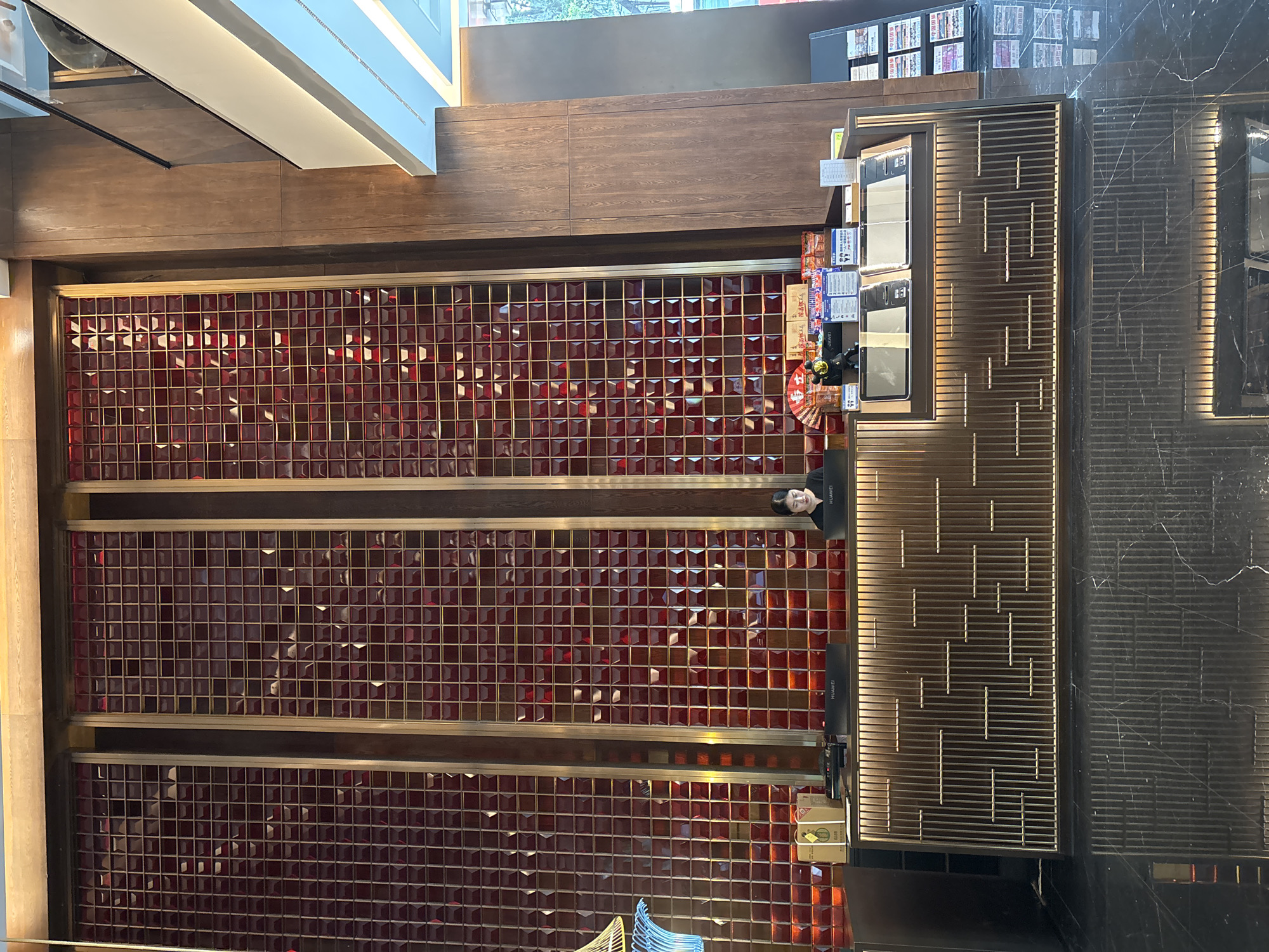 The image depicts the interior of a business, possibly a retail store, located in Liangjiang, China. The view is from above, looking down at a display wall.
The wall is composed of three sections, each filled with a grid of small, red, geometric tiles. These sections are separated by horizontal wooden panels.
To the right of the red-tiled wall is a counter or service area. There are several electronic devices, possibly point-of-sale systems or information displays, visible on the counter. Various items, including what appears to be promotional materials and packaged goods, are also arranged on or near the counter.
A person is standing near the service area, partially obscured by the counter and promotional materials. The person's attire and actions are not clearly discernible from the given perspective.
The environment appears modern and well-maintained. The floor is made of dark marble tiles. The overall impression is that this is a commercial establishment, possibly a shop selling local products or souvenirs.