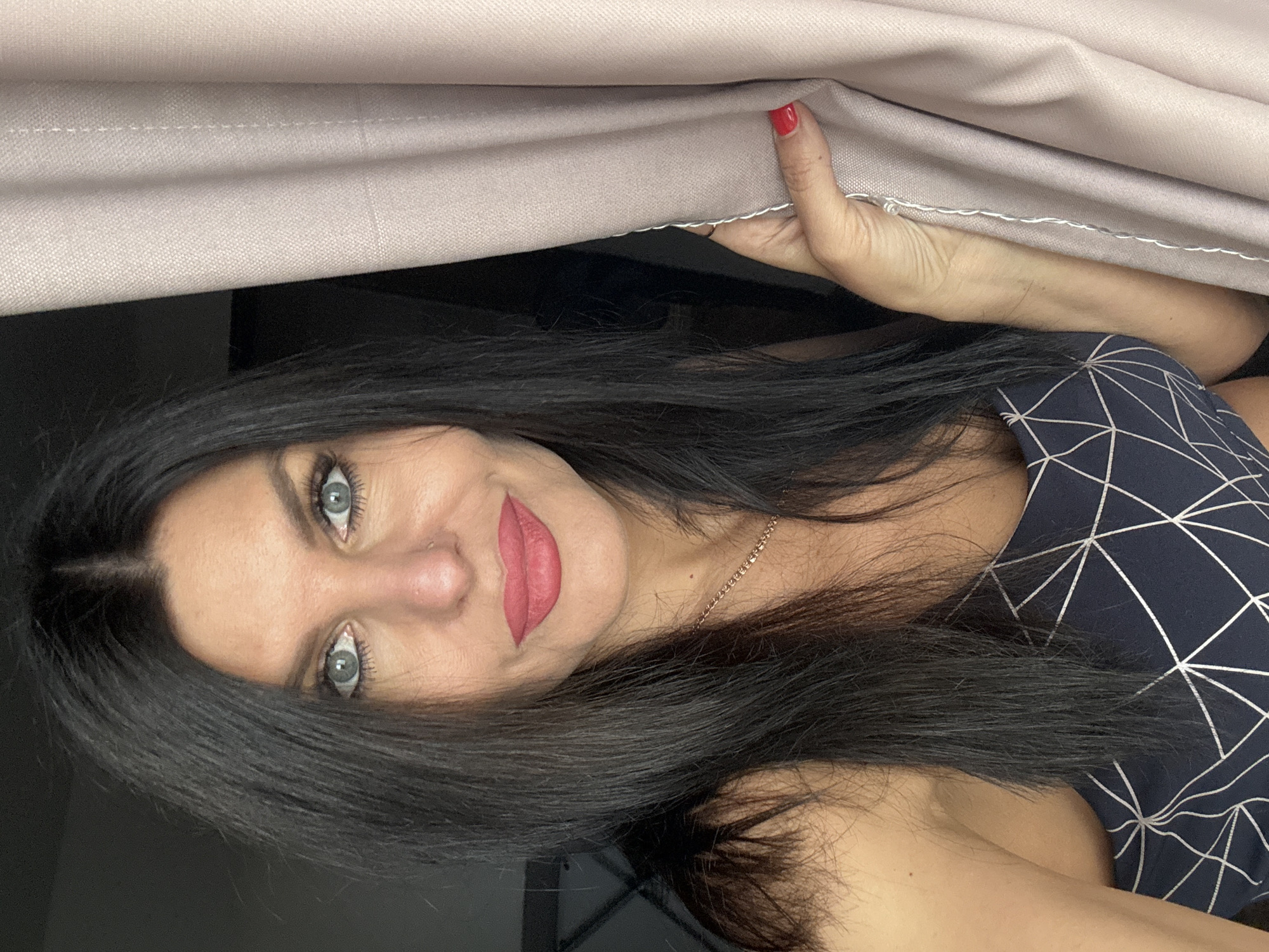 In a dimly lit indoor setting, a woman with long, dark hair and blue eyes, wearing a dark patterned top, poses by a light pink curtain. Her lips are painted a vibrant pink. She is looking directly at the camera. Her right hand, with red nail polish, is holding the curtain. The background is dark and indistinct. There is no visual information in the image that would suggest a location in Chaikovsky, Russia.