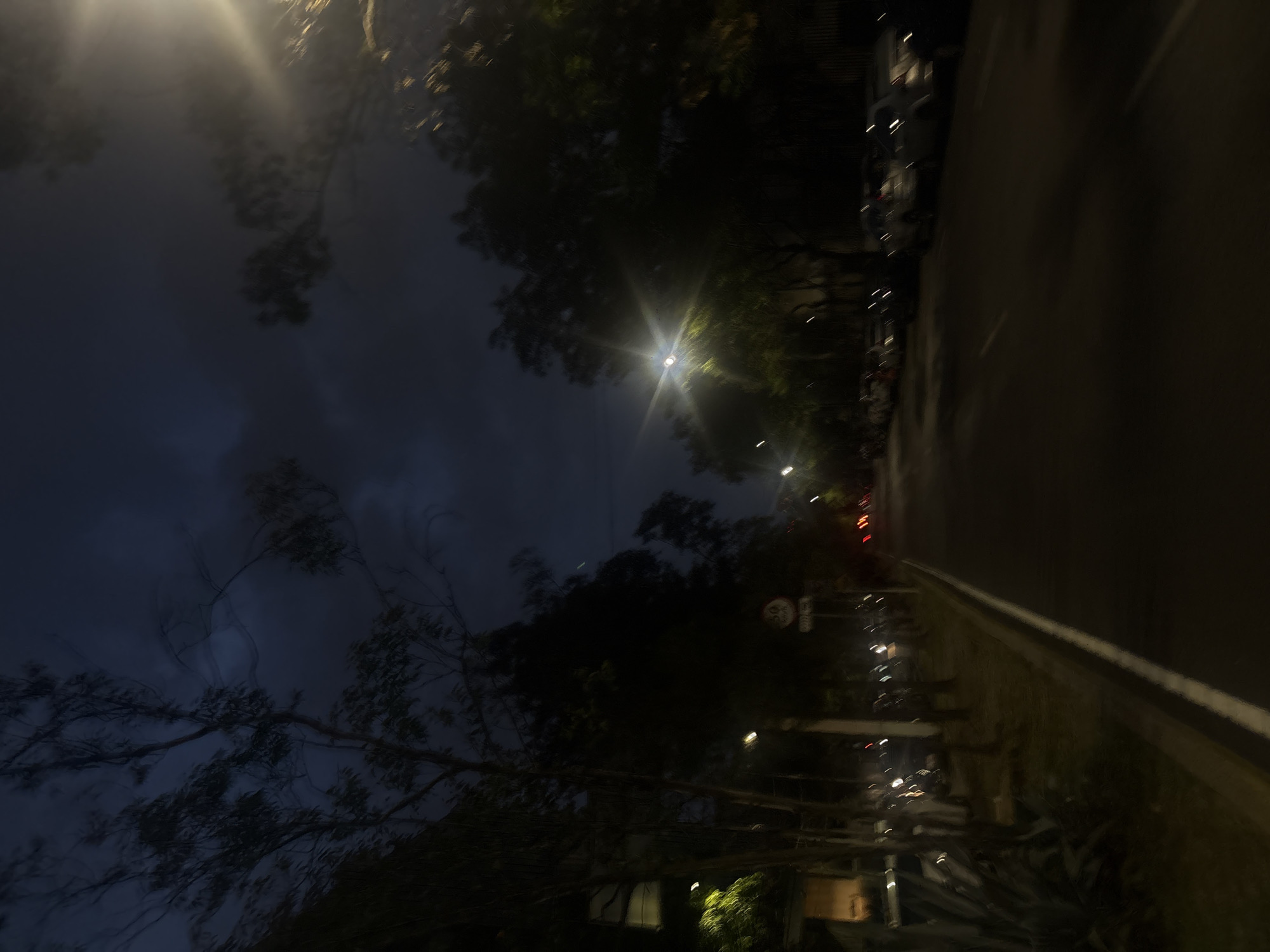 The image shows an outdoor scene at night. A dark asphalt road stretches into the distance, with parked cars lining the left side of the road. A street light illuminates the area. Trees are visible along the roadside. The photo was taken in Mexico City, Mexico.