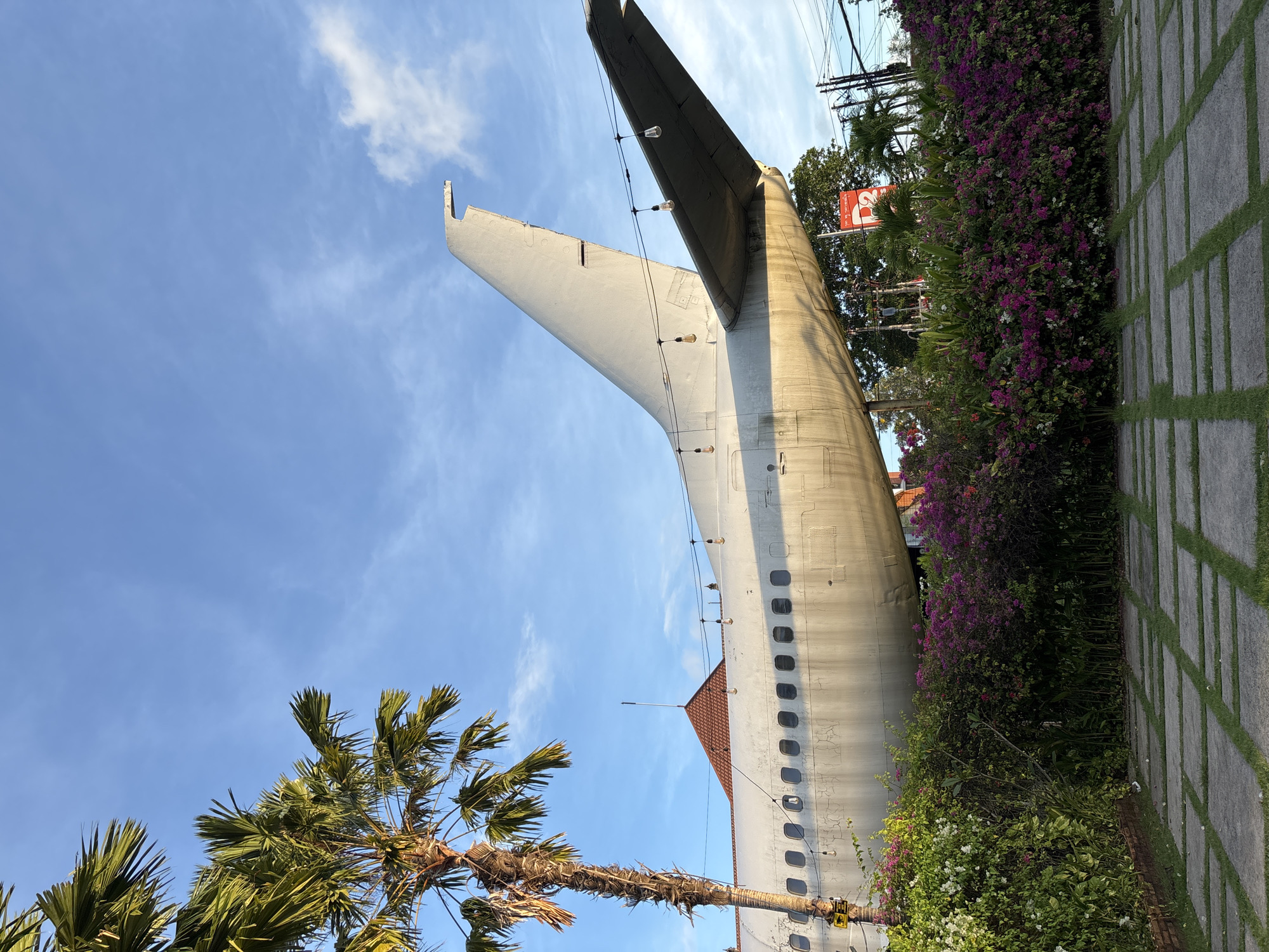 The image depicts the tail section of an aircraft in Kuta, Indonesia, seemingly repurposed as part of a building structure or an unusual architectural feature. The tail is white, and displays the tail fin and horizontal stabilizer. There are a string of lights alongside the length of the tail. The plane is surrounded by greenery, including a palm tree on the lower left and flowering bushes on the right. A tiled walkway with grass growing between the tiles is on the right side of the frame. The sky is blue with scattered clouds. A red-and-white sign is seen through the vegetation.