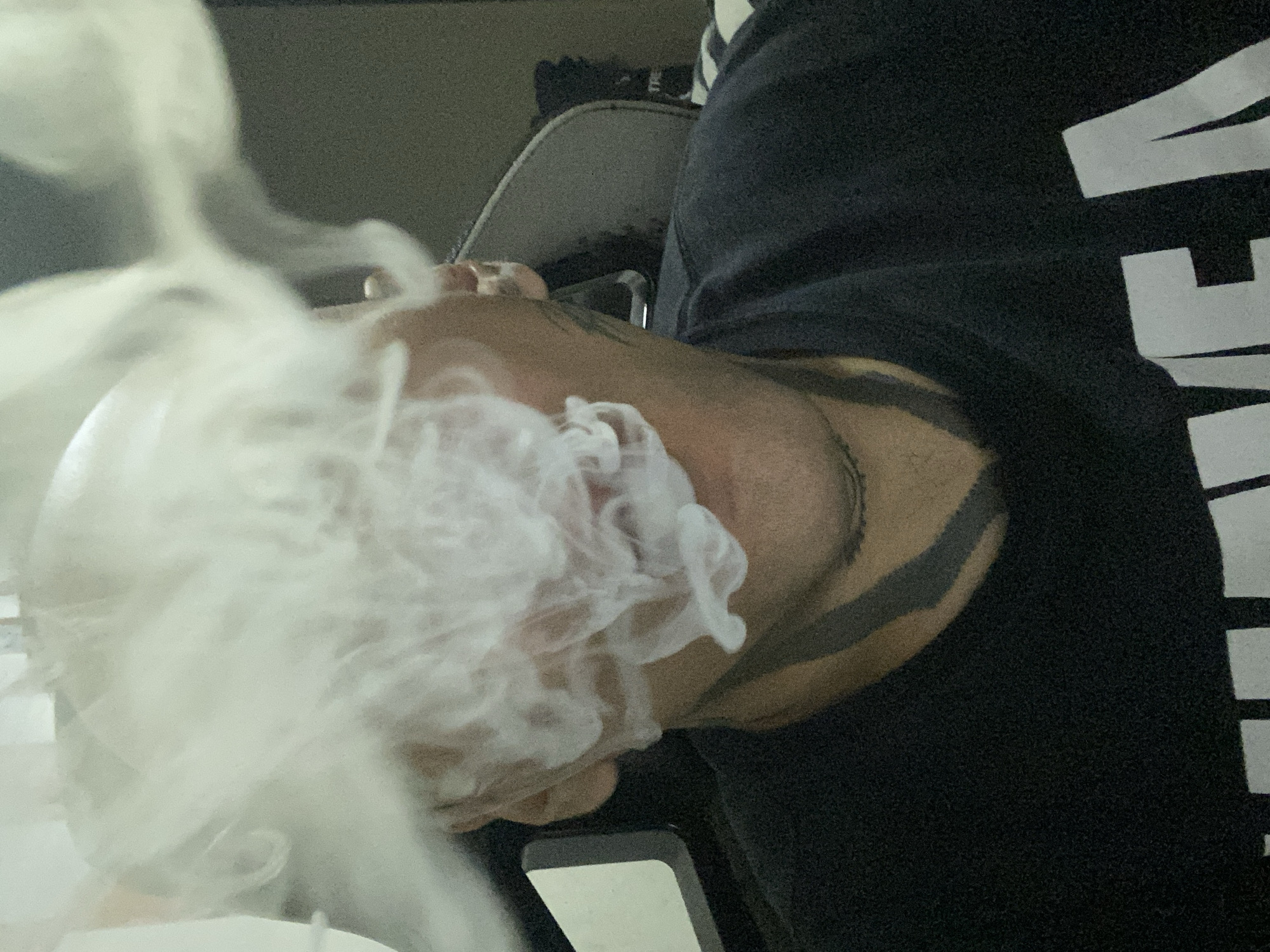 A close-up shot shows a person exhaling a cloud of white smoke. The person has a bald head and dark tattoos visible on their neck and face. They are wearing a black hooded sweatshirt with white lettering. The background is indistinct but includes a light-colored chair and wall. There is no information provided in the image to confirm that this is Buenos Aires, Argentina.