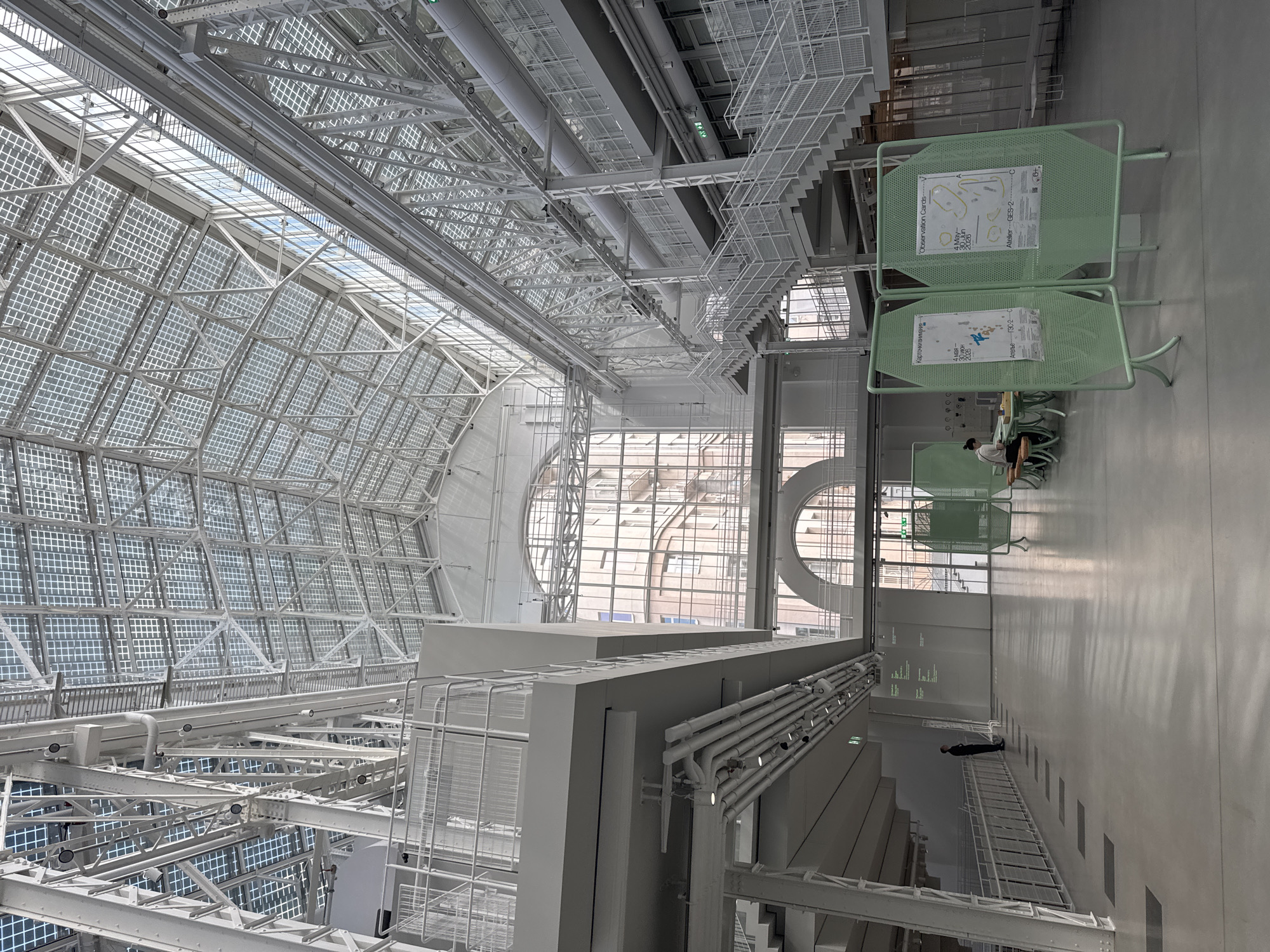 A high-angle, vertically oriented photograph captures the vast, modern interior of a building, likely the GES-2 House of Culture in Moscow, Russia. The space is characterized by an expansive, angular glass and metal roof structure that dominates the upper portion of the image. This roof, composed of numerous square glass panels within a white grid framework, allows abundant natural light to flood the interior. Below the roof, intricate white metal trusses, ventilation ducts, pipes, and lighting fixtures are visible, creating an industrial yet refined aesthetic.
On the ground level, a polished light-colored floor stretches into the distance. A few individuals are present, adding scale to the grand space. One person is seated at a table near two prominent light-green, perforated metal stands, while another stands further down the hall. The green stands display information, including both Russian and English text.
Visible text on the stands includes "Observation Cards" in both Russian ("Карточкивнимания") and English, indicating a temporary exhibition or interactive element. Dates "4 May - 30 Jun 2026" are clearly displayed, suggesting the period of this event. The text "Atelier—GES-2-" also appears, explicitly linking the location to the GES-2 House of Culture and possibly an associated workshop or studio. Large, arched windows in the background offer glimpses of the exterior. The overall impression is one of modern architecture, spaciousness, and bright, natural illumination, with minimal human activity in the captured moment.