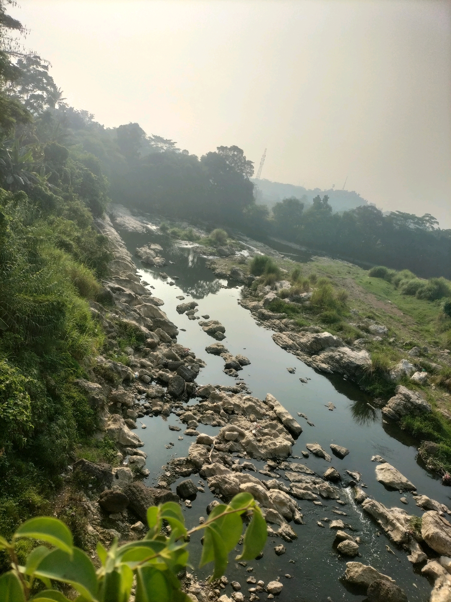 The image shows a rocky river flowing through a natural landscape. The river is partially filled with rocks, with some flowing water visible. The banks of the river are covered in green vegetation and trees. In the background, there are hills and a hazy sky, with two construction cranes visible on the horizon. The scene appears to be an outdoor natural setting, possibly a park or a rural area, with some signs of ongoing construction in the distance. The lighting suggests it is daytime, possibly early morning or late afternoon, given the hazy atmosphere.