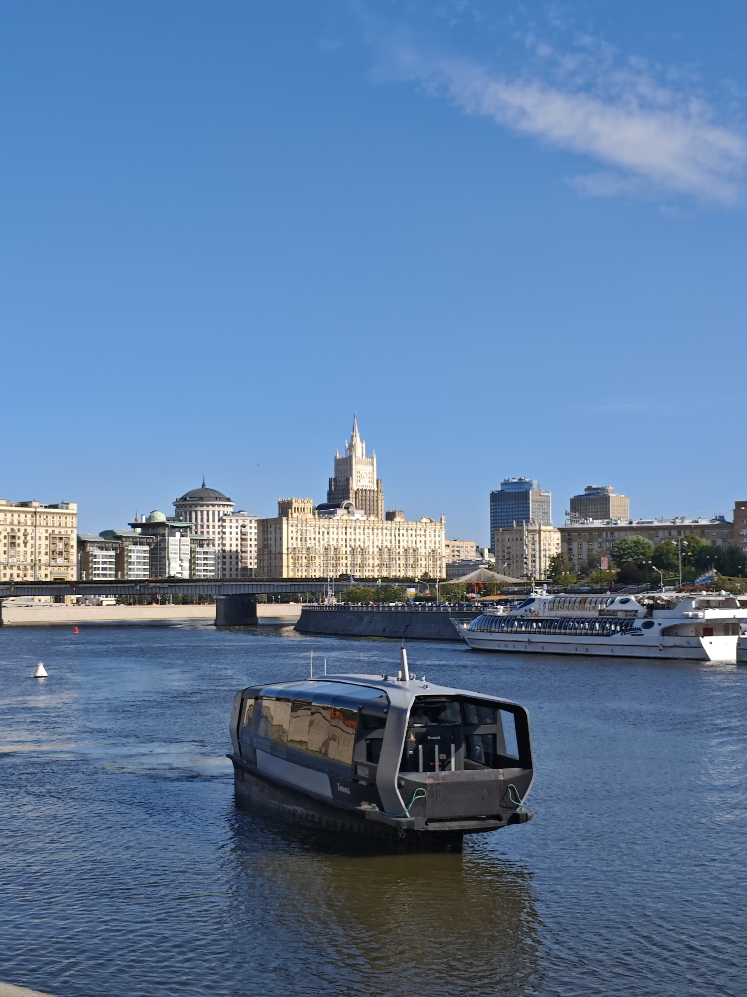A modern, dark-colored passenger boat with a glass enclosure is on the Moscow River, its stern pointed towards the viewer. In the background, across the river, a large, ornate building with a spire dominates the skyline, flanked by other buildings and a bridge. A larger, white passenger ship is also visible on the river to the right of the modern boat. The sky is clear blue with a few wispy clouds. The scene appears to be in Moscow, Russia.