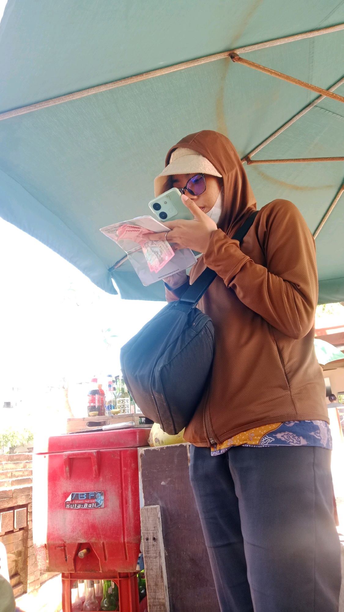 A person wearing a brown hooded jacket, a light-colored bucket hat, and sunglasses is looking at a mobile phone while holding several banknotes. They have a black bag slung over their shoulder. In the background, a red cooler with a label reading "IB.F BEER FACTORY KUTA-BALI" is visible, along with several bottles. The scene appears to be outdoors, possibly a street stall in Kuta, Indonesia, with a large, light blue umbrella overhead providing shade. The lighting suggests it is daytime.