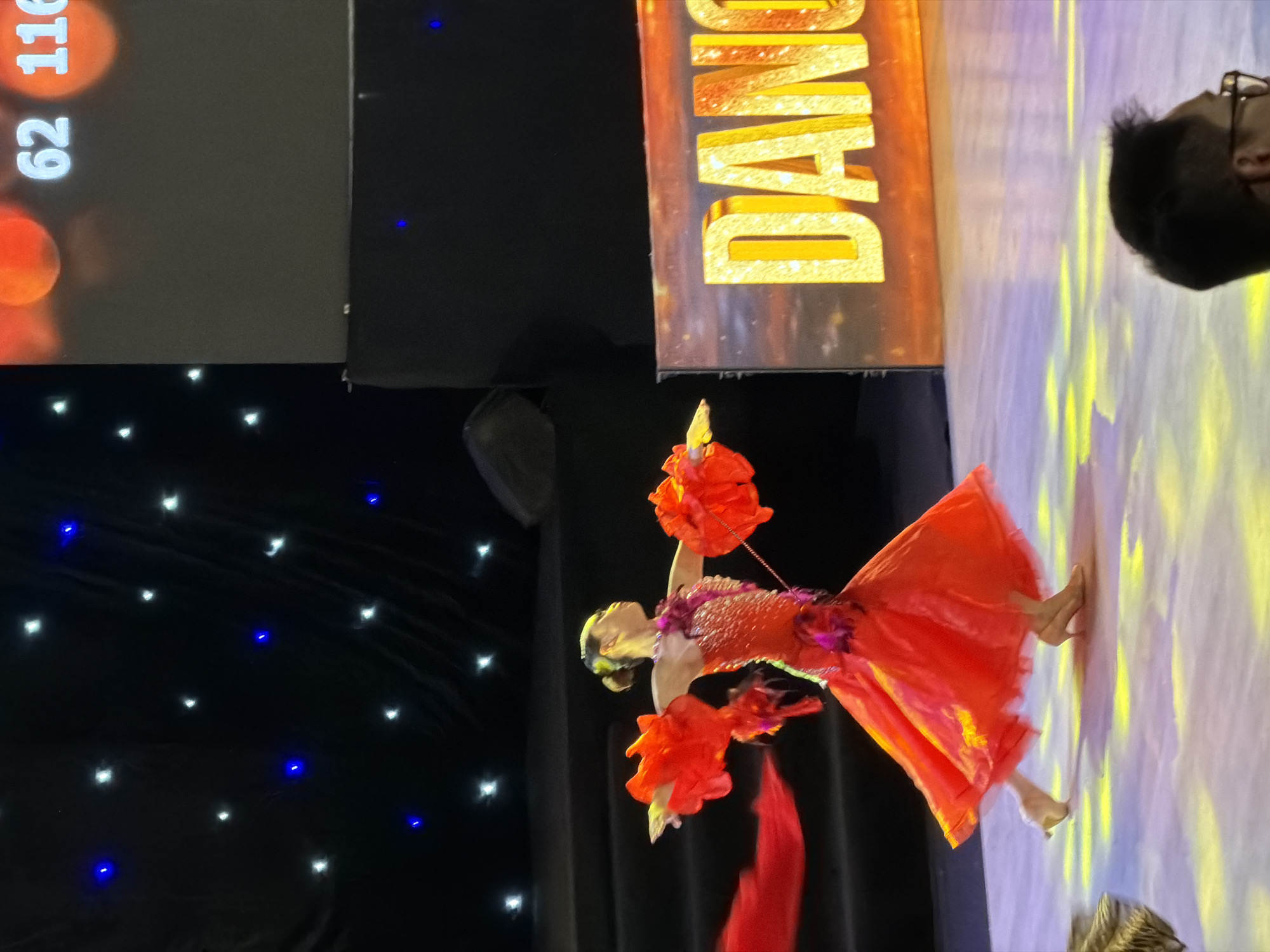 A female dancer in a vibrant red, ornate dress with exaggerated sleeves is captured mid-performance on a brightly lit stage. The dress features intricate beadwork and floral embellishments. Her arms are extended, with one hand holding a flowing red ribbon. Her hair is pulled back in a bun. The stage floor is illuminated with yellow and purple lights. In the background, a large screen displays the word "DANCE" in bold, sparkling yellow letters against a backdrop of an orange and brown sunset. To the left of the dancer, a dark backdrop is adorned with small blue and white lights, resembling stars. A digital scoreboard with numbers "62" and "116" is visible at the top right of the screen. The scene appears to be from a dance competition or performance, potentially within a venue in Hà Nội, Vietnam, given the context. The lighting suggests a professional performance setting.